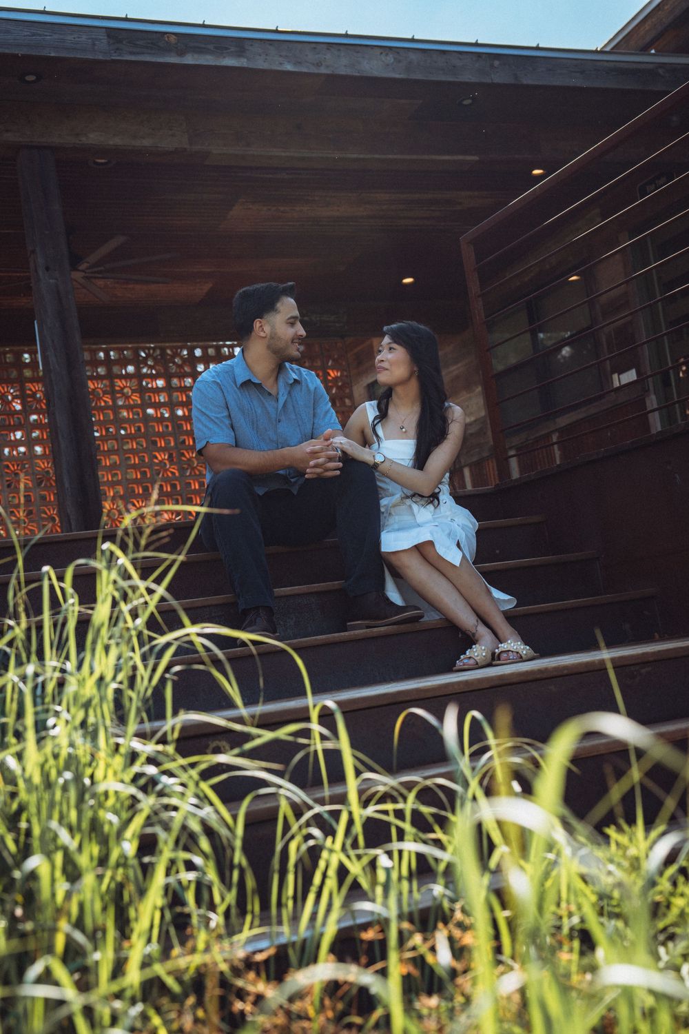 A couple sits on wooden steps, smiling at each other, surrounded by lush green grass near a rustic building.