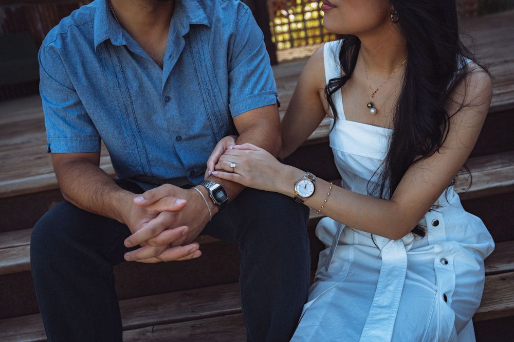 Couple sitting on wooden steps holding hands, woman in white dress with watch, man in blue shirt.