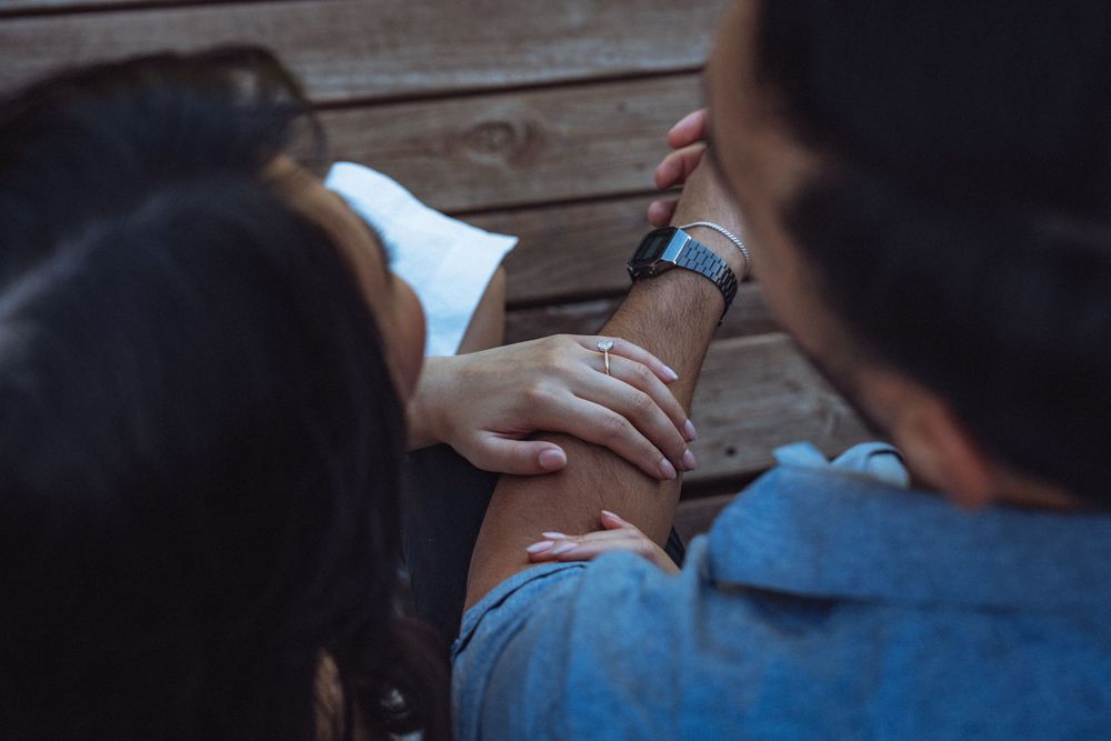 Two people sitting closely together on wooden steps, one resting hand on the other's arm wearing a smartwatch.