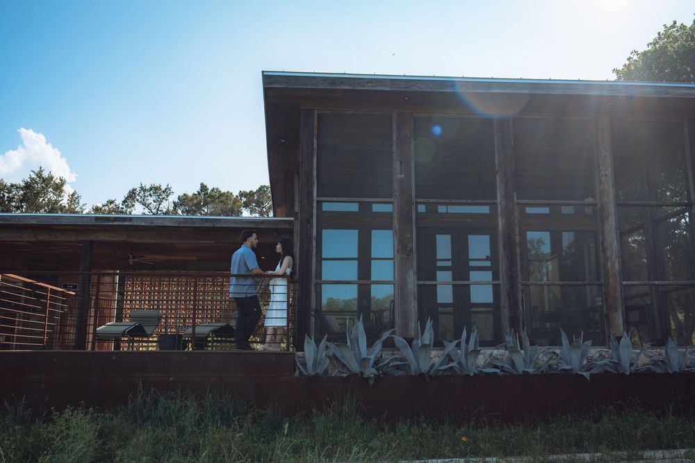 Couple standing on a rustic wooden porch of a modern farmhouse building under a bright blue sky.