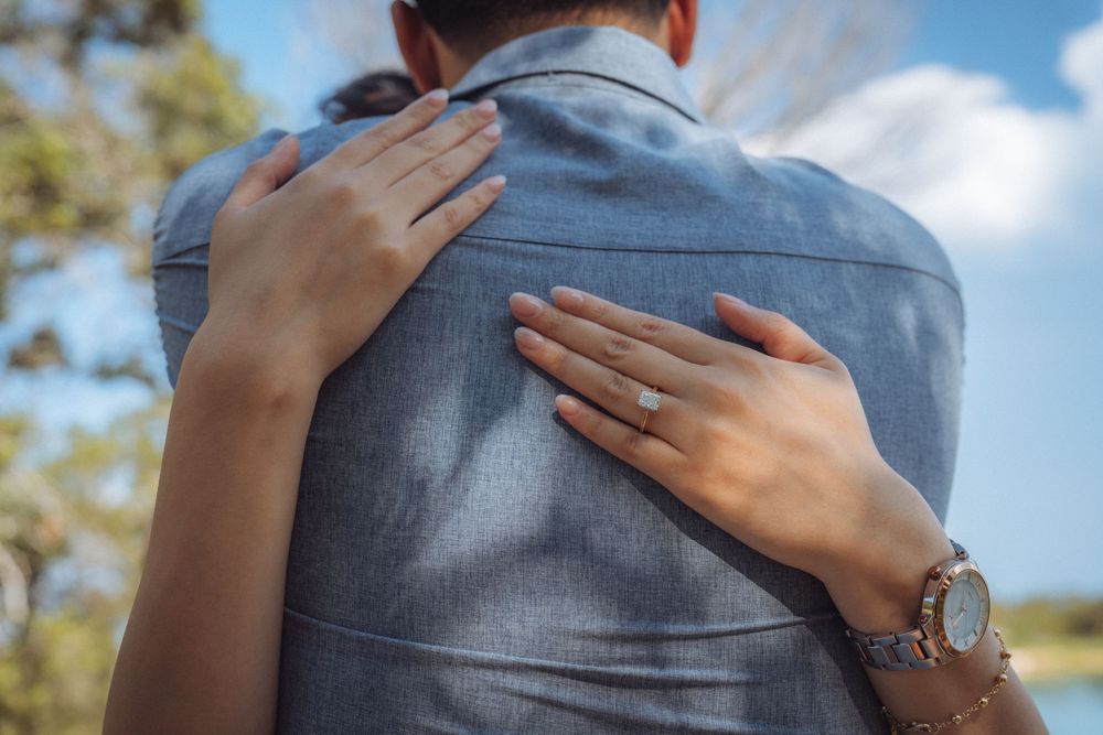 A woman with an engagement ring and watch embraces a man in a gray shirt outdoors near trees and blue sky.