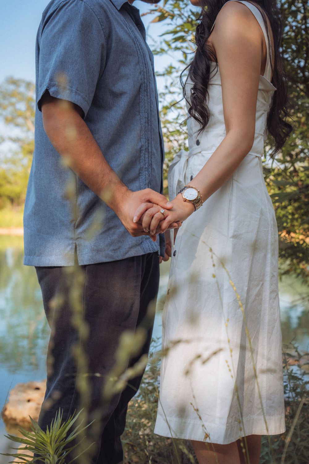 Couple holding hands outdoors near a lake, woman in white dress with watch, man in blue shirt, surrounded by greenery.