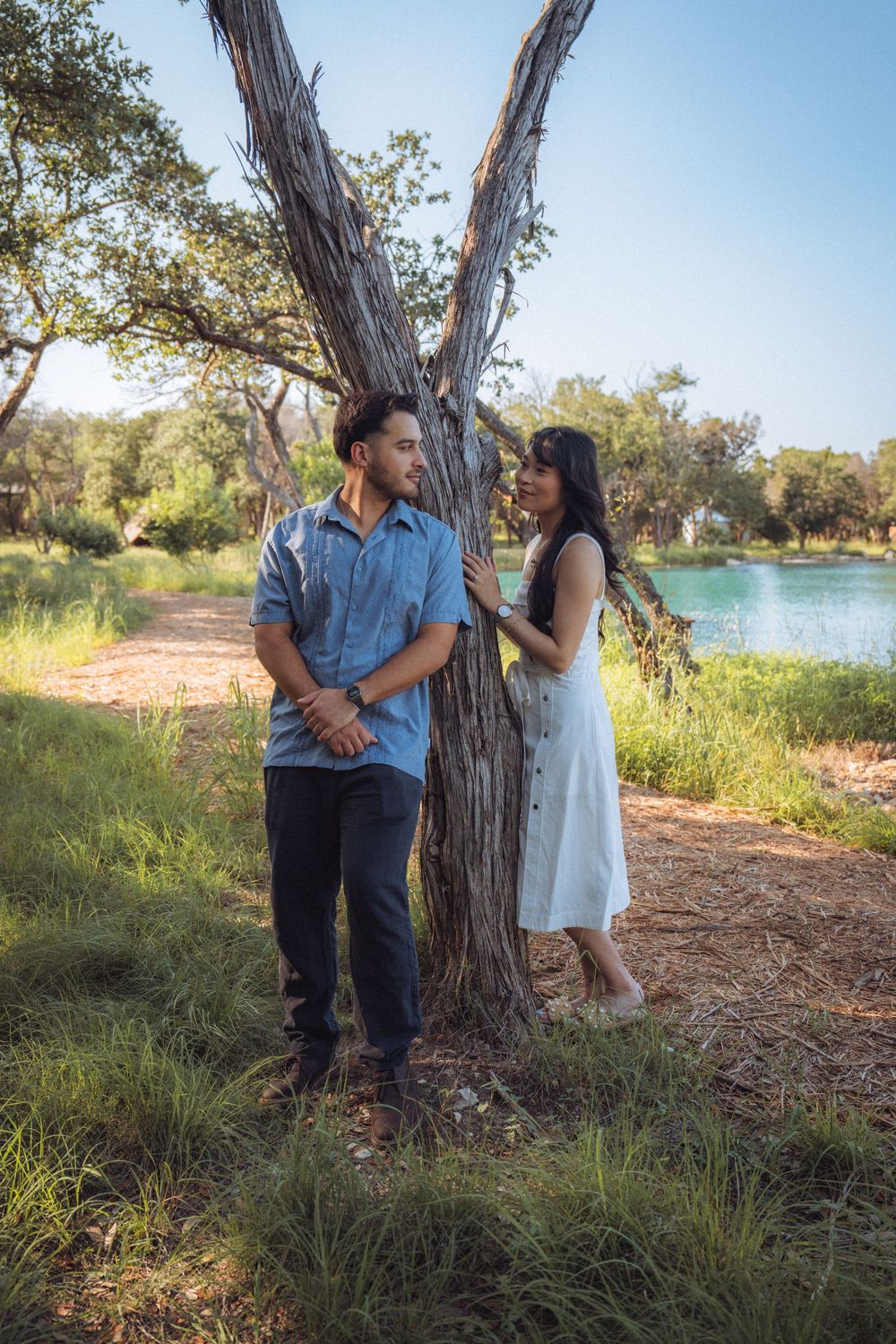 A couple stands by a large tree near a lake, the woman in white touching the tree while the man in blue looks away.