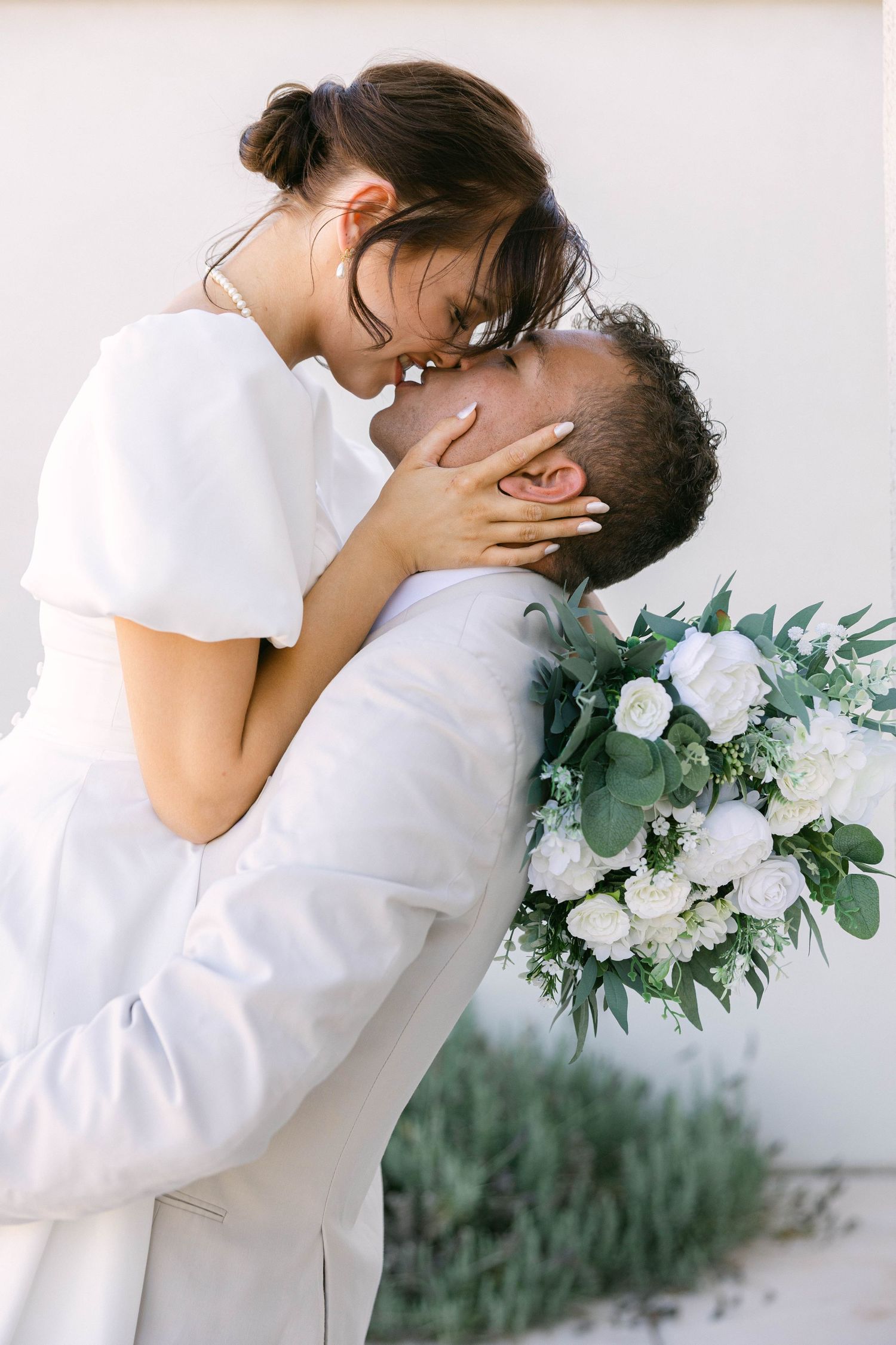 A bride and groom share a romantic kiss, with the bride holding a white floral bouquet with greenery on their wedding day.