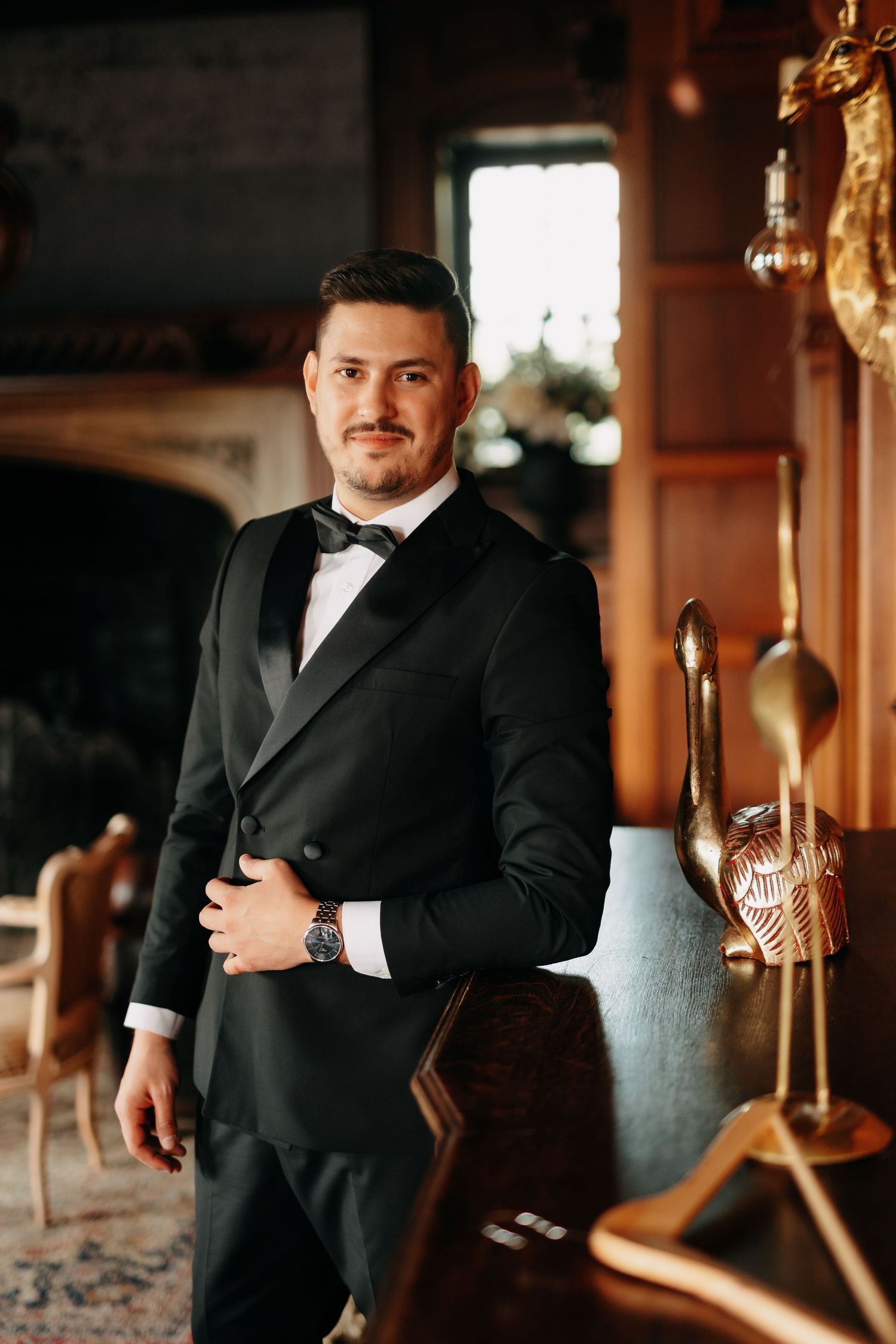 Elegant man in black tuxedo with bow tie and watch posing in a luxurious wood-paneled room.