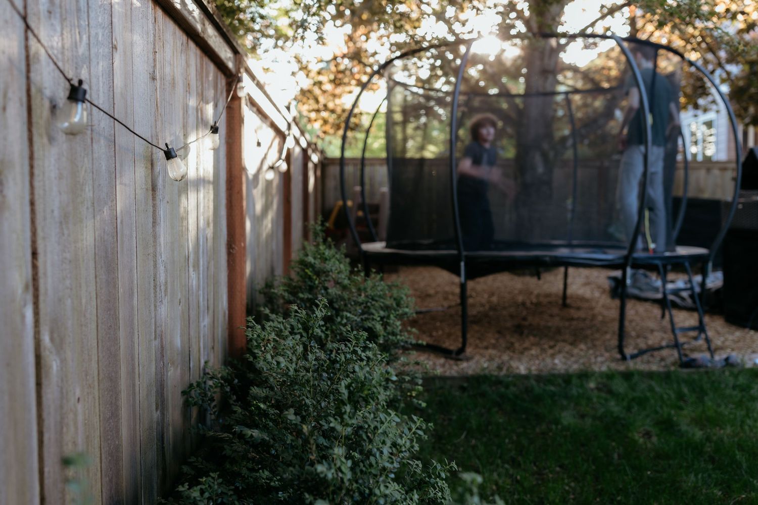 Child playing on a backyard trampoline near a wooden fence with string lights and lush green shrubs.