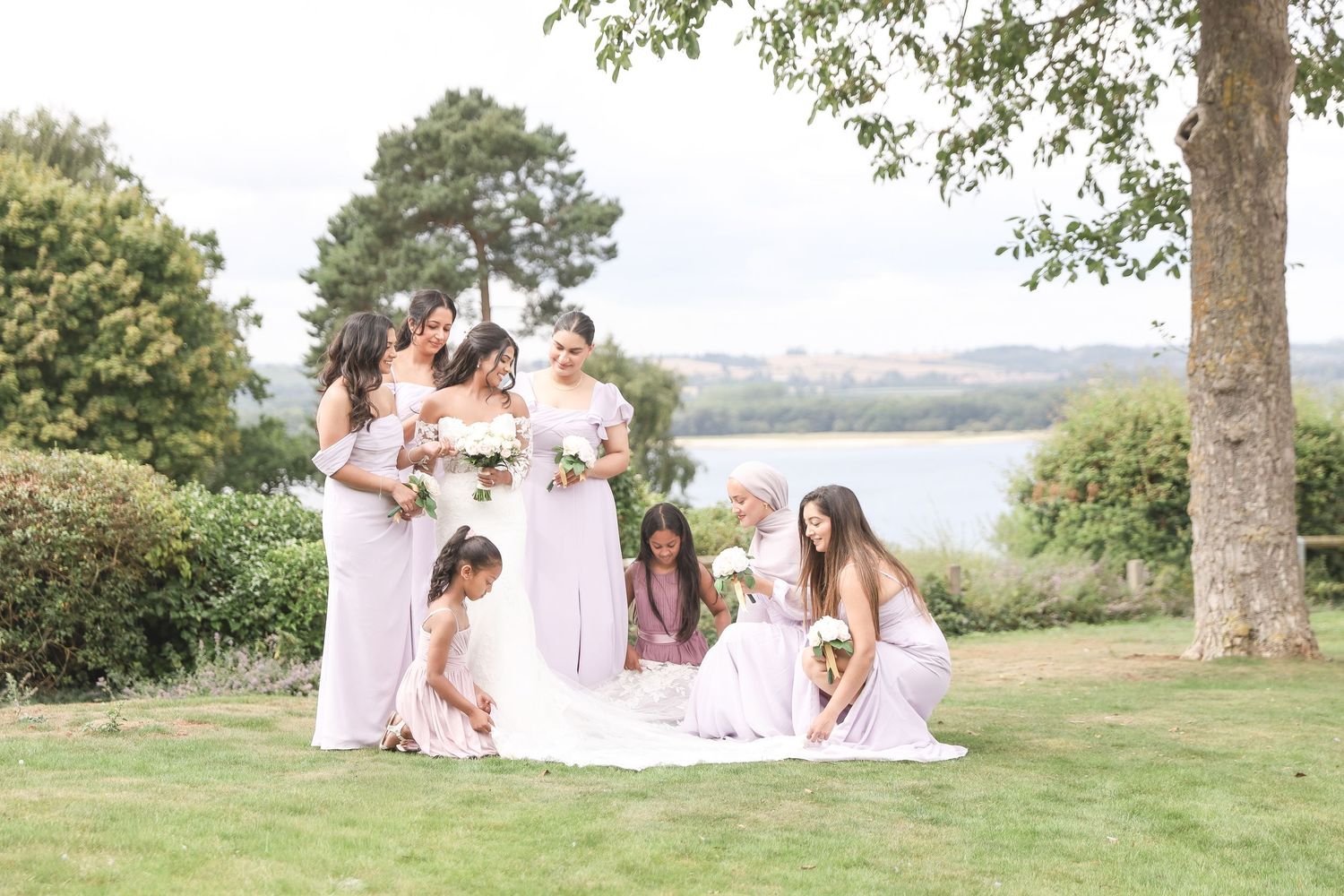 Bridesmaids in blush pink gowns gather around a bride on a lush green lawn near a lake on a sunny wedding day.