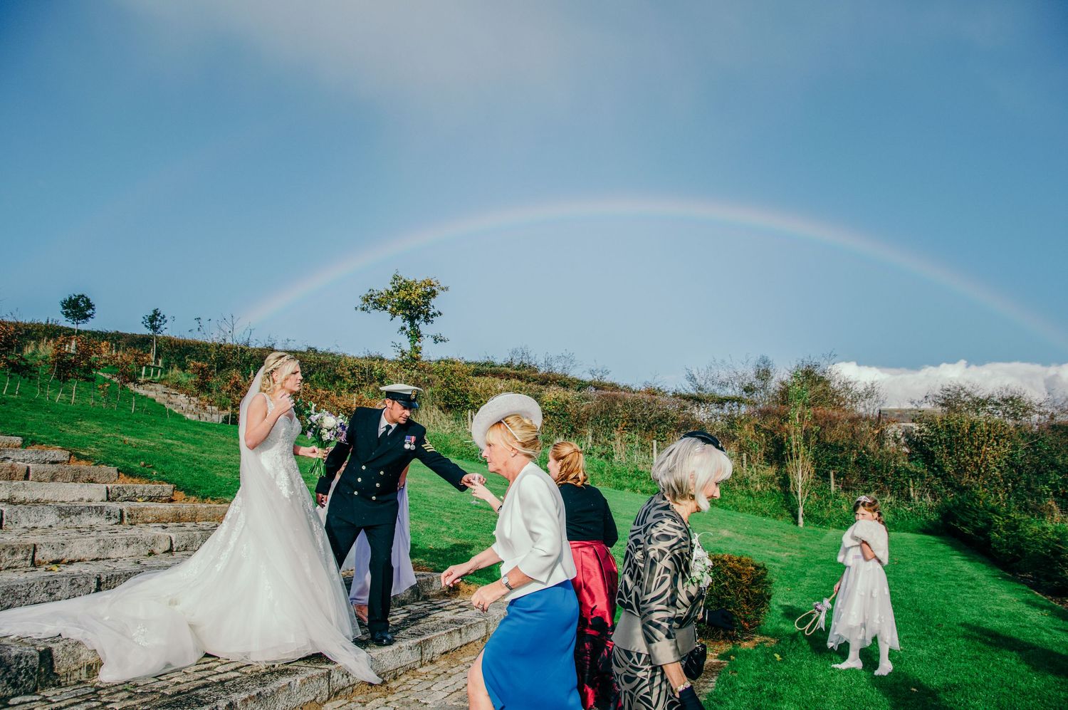Wedding party celebrates on stone steps under a stunning rainbow on a sunny day at an outdoor venue.