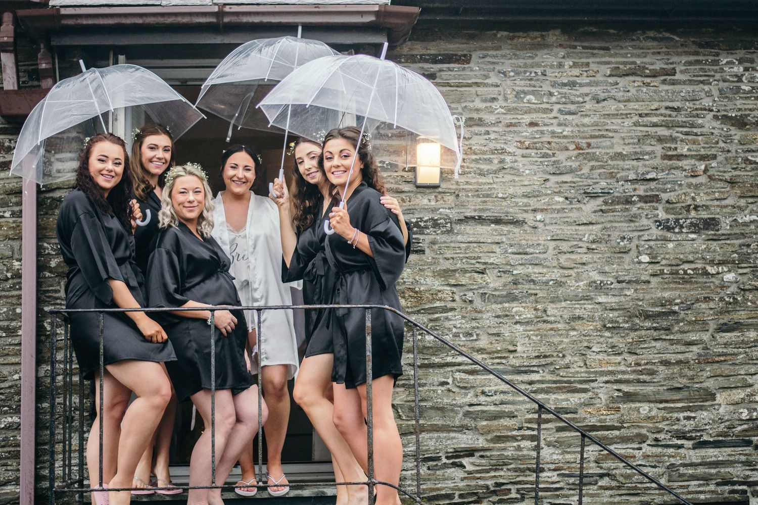 Bridesmaids in black robes holding clear umbrellas pose with bride on rainy day outside stone building.