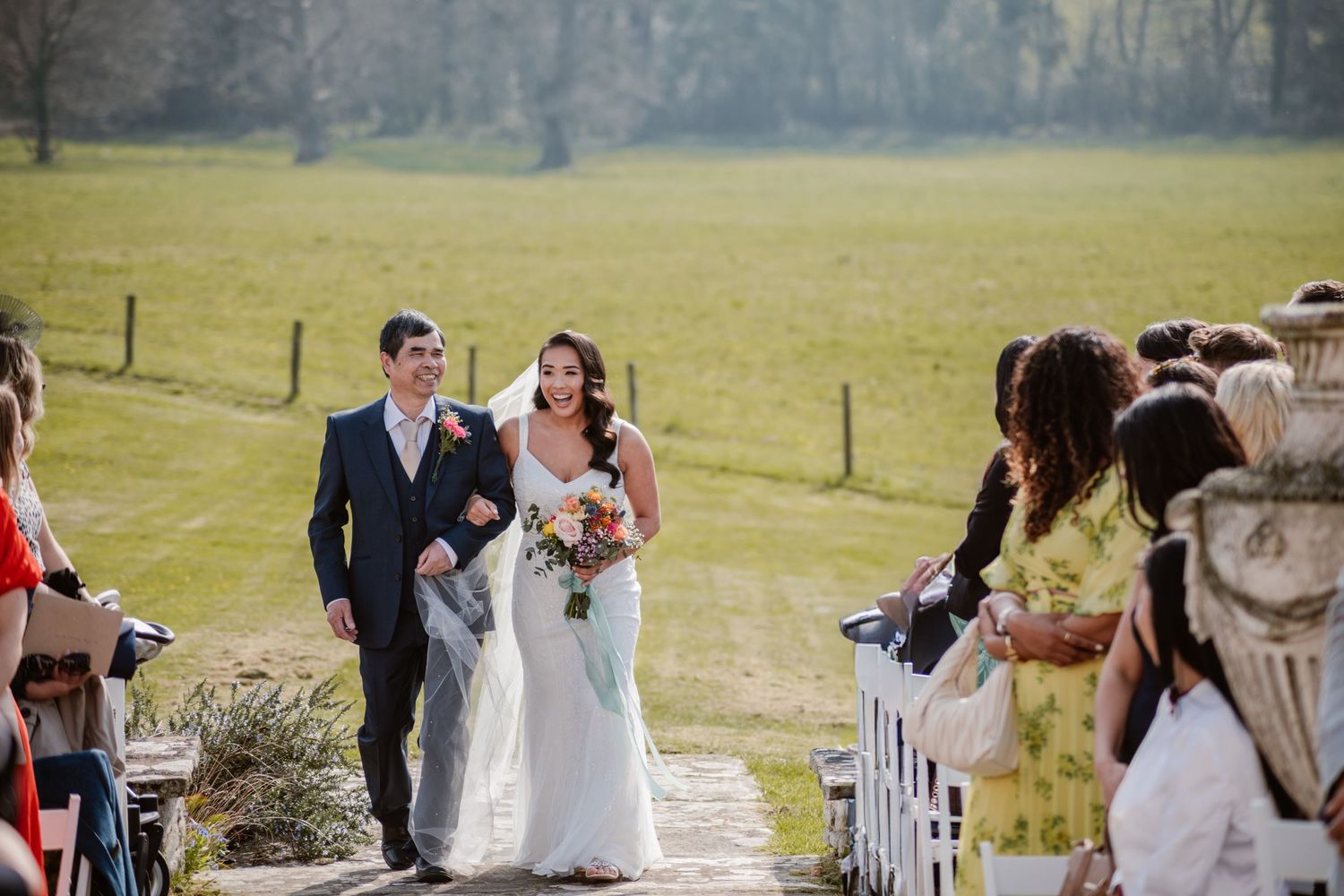 Groom in blue suit lifts laughing bride in white gown before vibrant pink rhododendron blooms in garden.