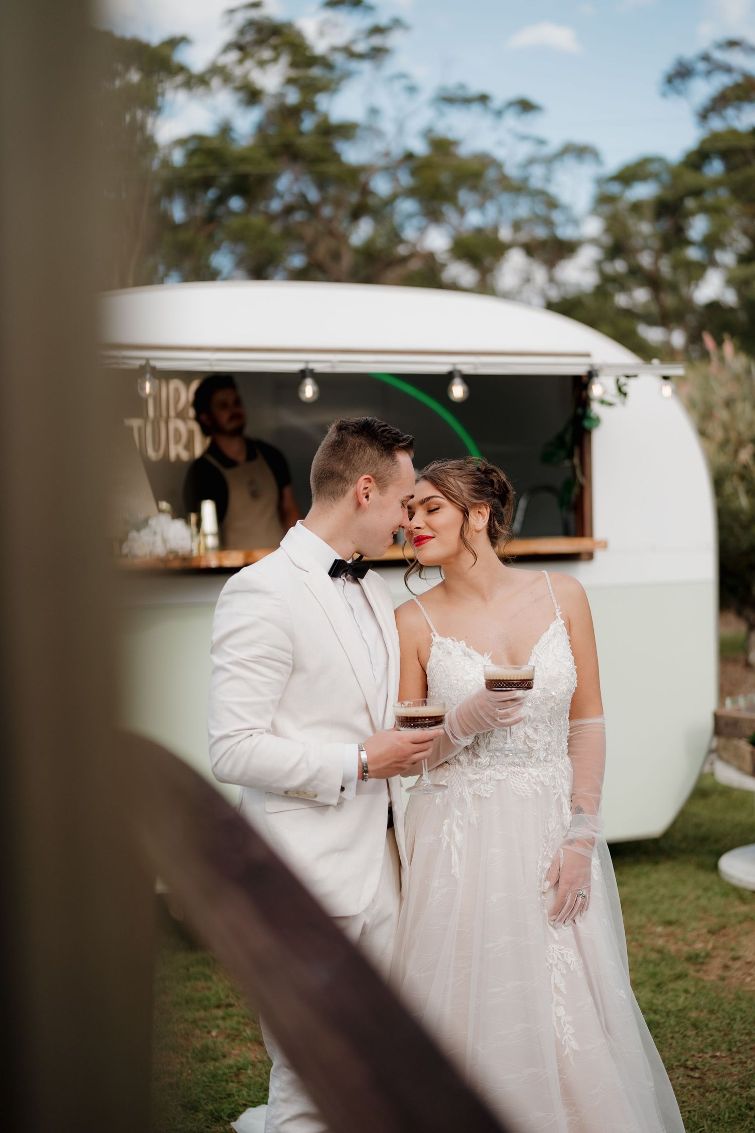 Bride and groom share a romantic moment holding drinks in front of a vintage white caravan bar at their outdoor wedding.