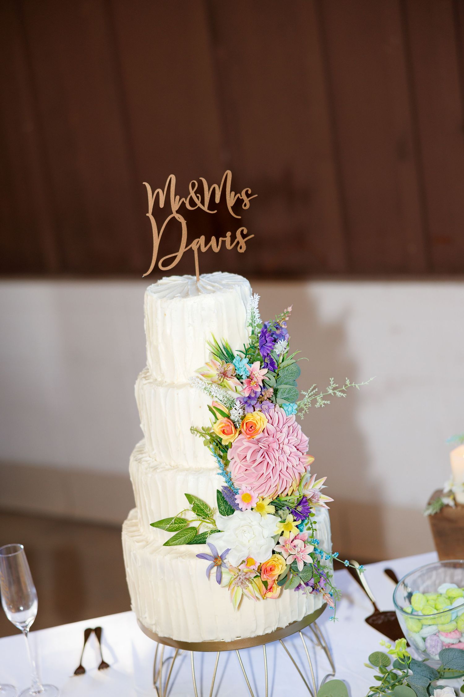 Elegant four-tier white wedding cake with colorful floral decorations and Mr & Mrs Davis cake topper on a gold stand.