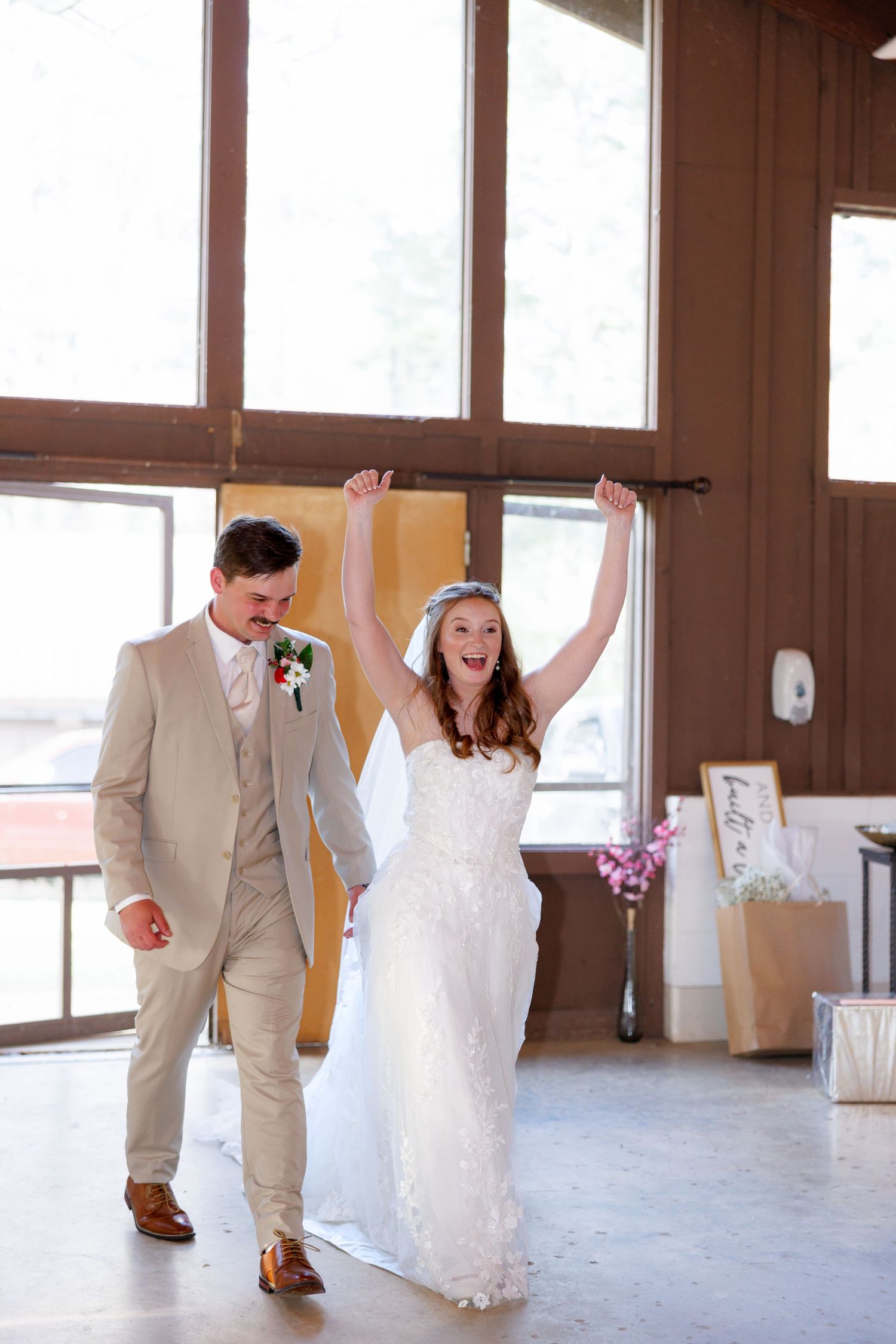 Joyful bride in white gown raises arms as groom in tan suit escorts her into rustic wedding reception hall.