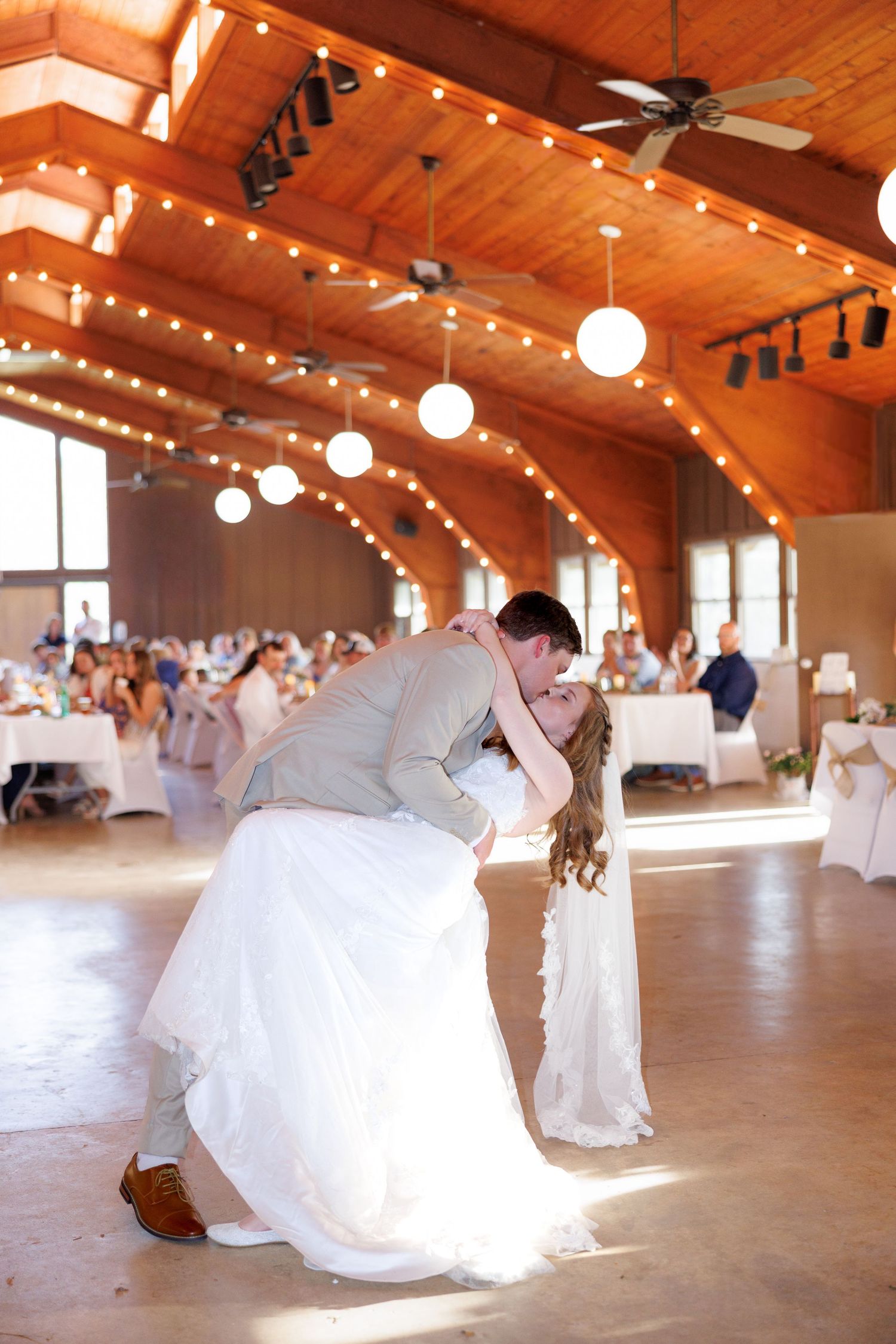 Bride and groom share a romantic dip kiss on the dance floor at a rustic wedding reception with string lights.