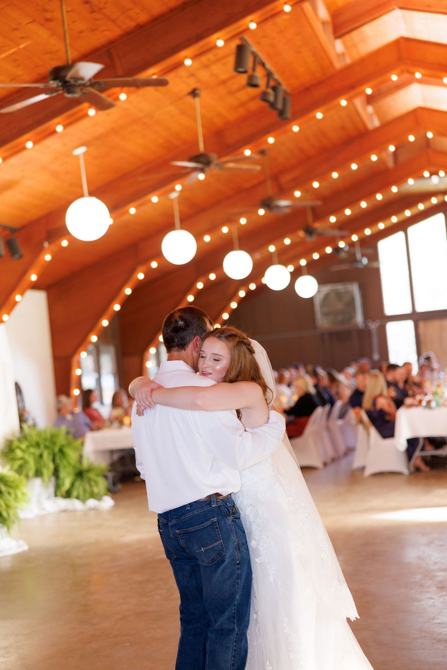 Bride and groom share first dance in elegant wood-beamed reception hall adorned with string lights and globe pendants.