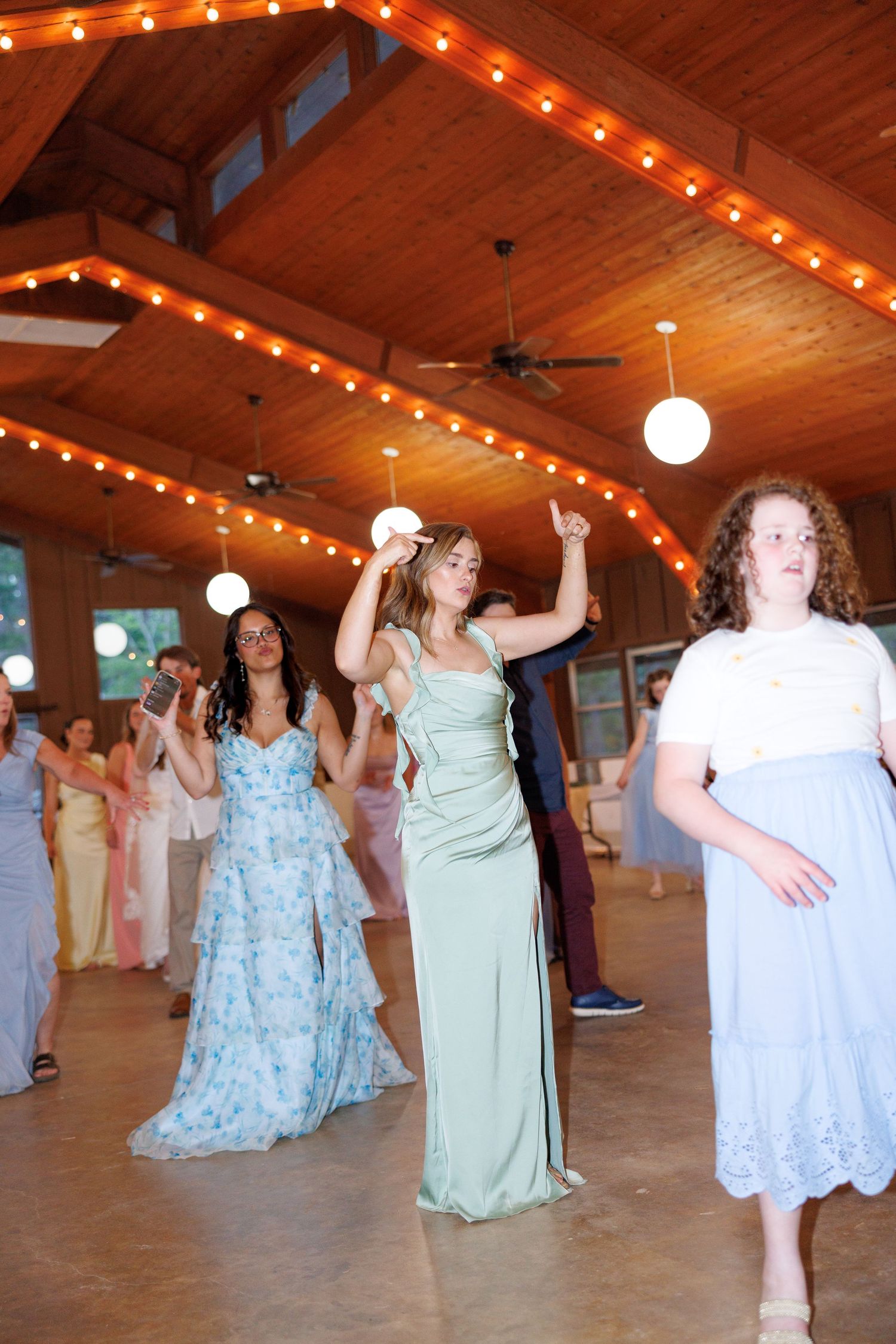 Guests in elegant pastel gowns dance at a rustic wooden venue decorated with warm string lights.