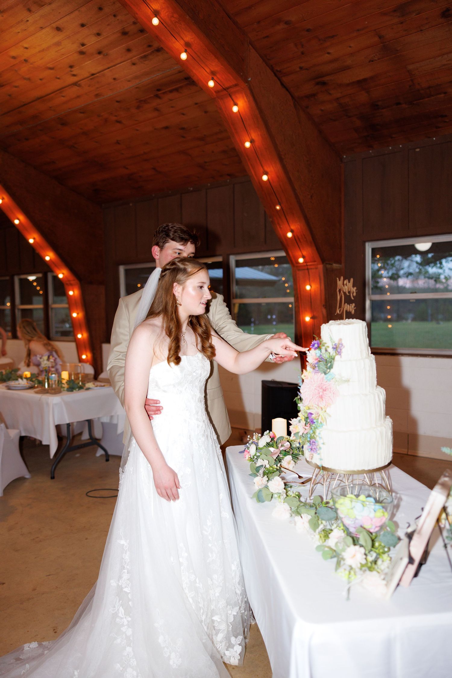 Bride and groom cutting a four-tier white wedding cake decorated with flowers in a rustic wood venue with string lights.