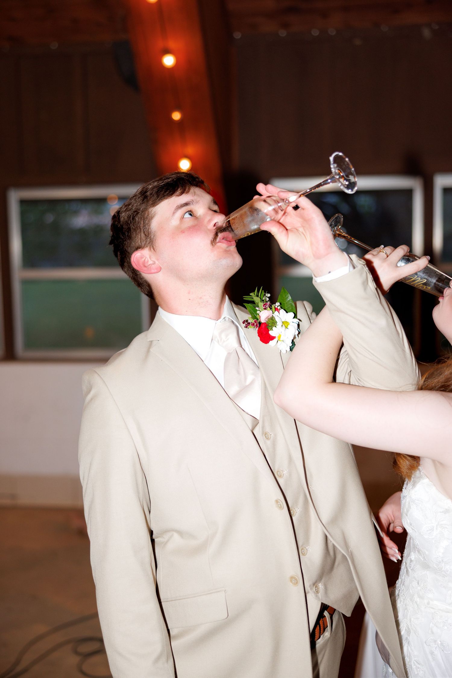 Groom in tan suit with boutonniere drinking champagne at wedding reception celebration.