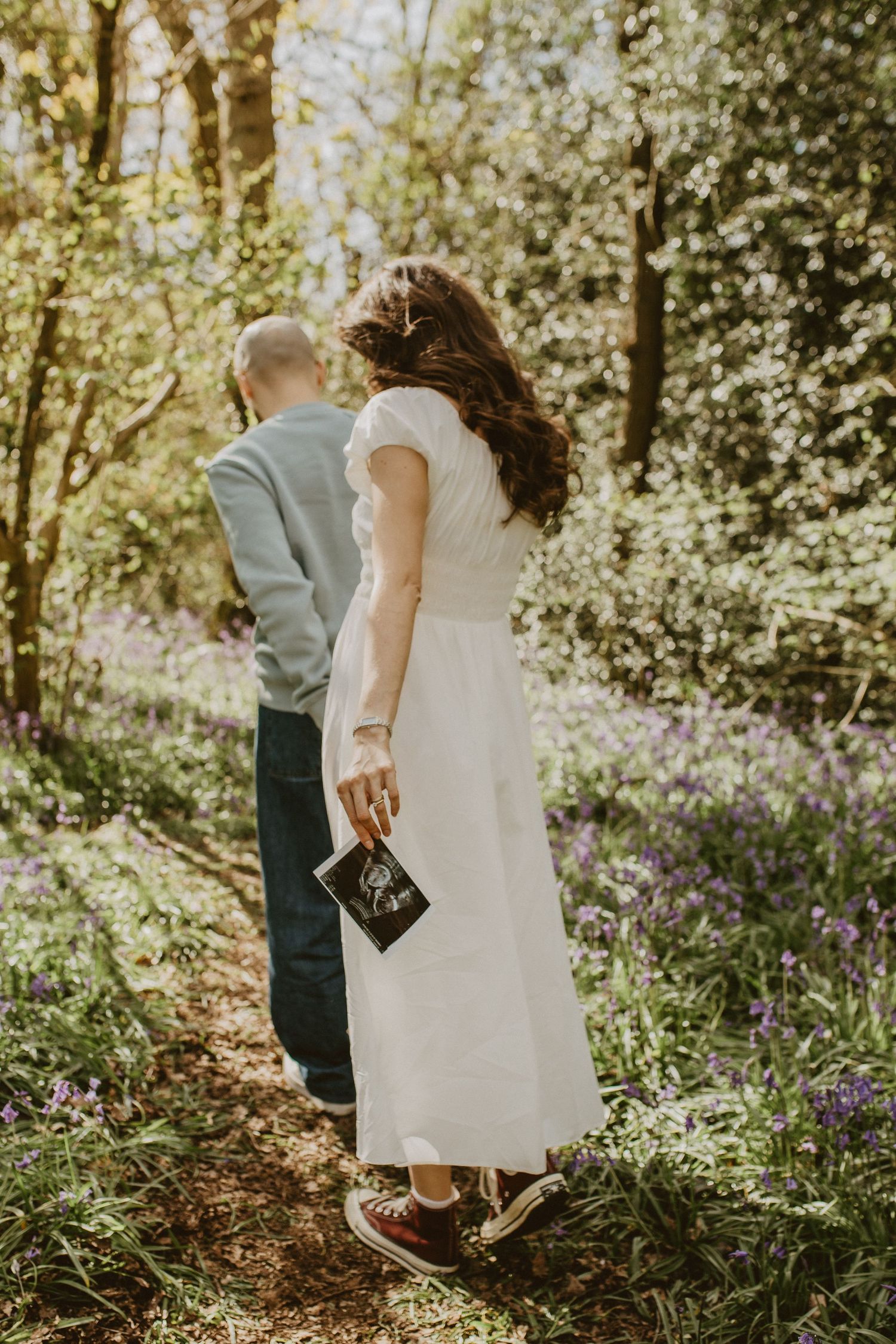 A couple walks through a bluebell woodland, the woman in a white dress holding an ultrasound scan photo.