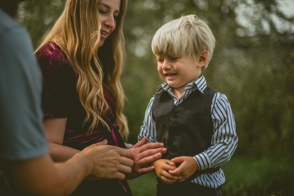 A young blonde boy in a striped shirt and vest smiles while interacting with a woman outdoors.