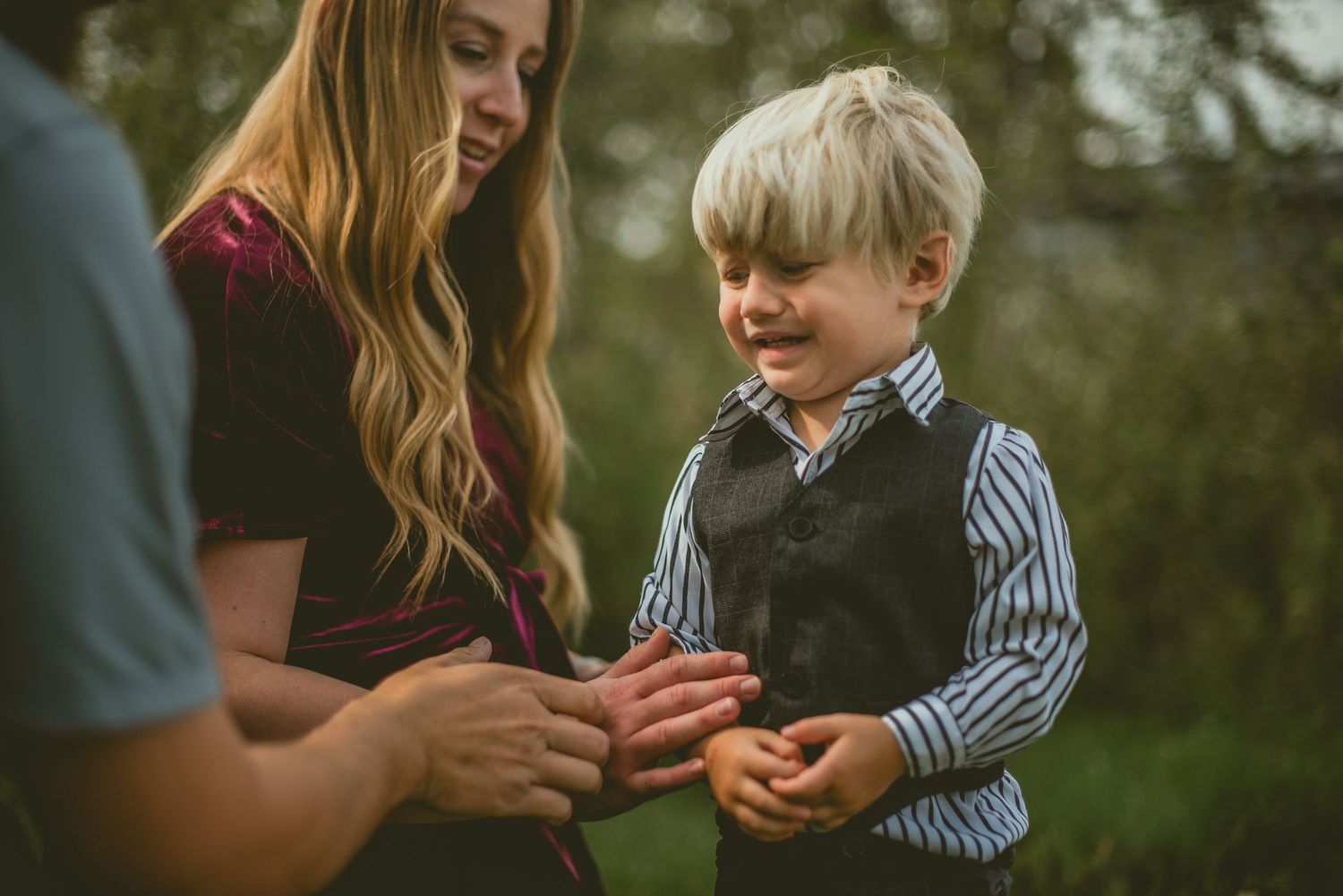 A young blonde boy in a striped shirt and vest smiles while interacting with a woman outdoors.