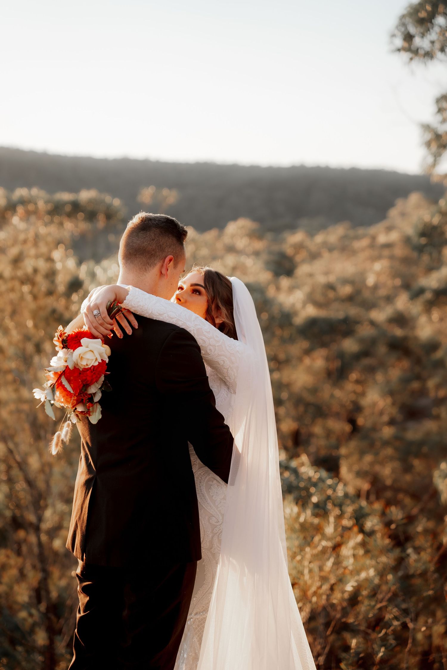 Bride and groom embracing outdoors with red and white bouquet, scenic autumn forest backdrop in warm sunlight.