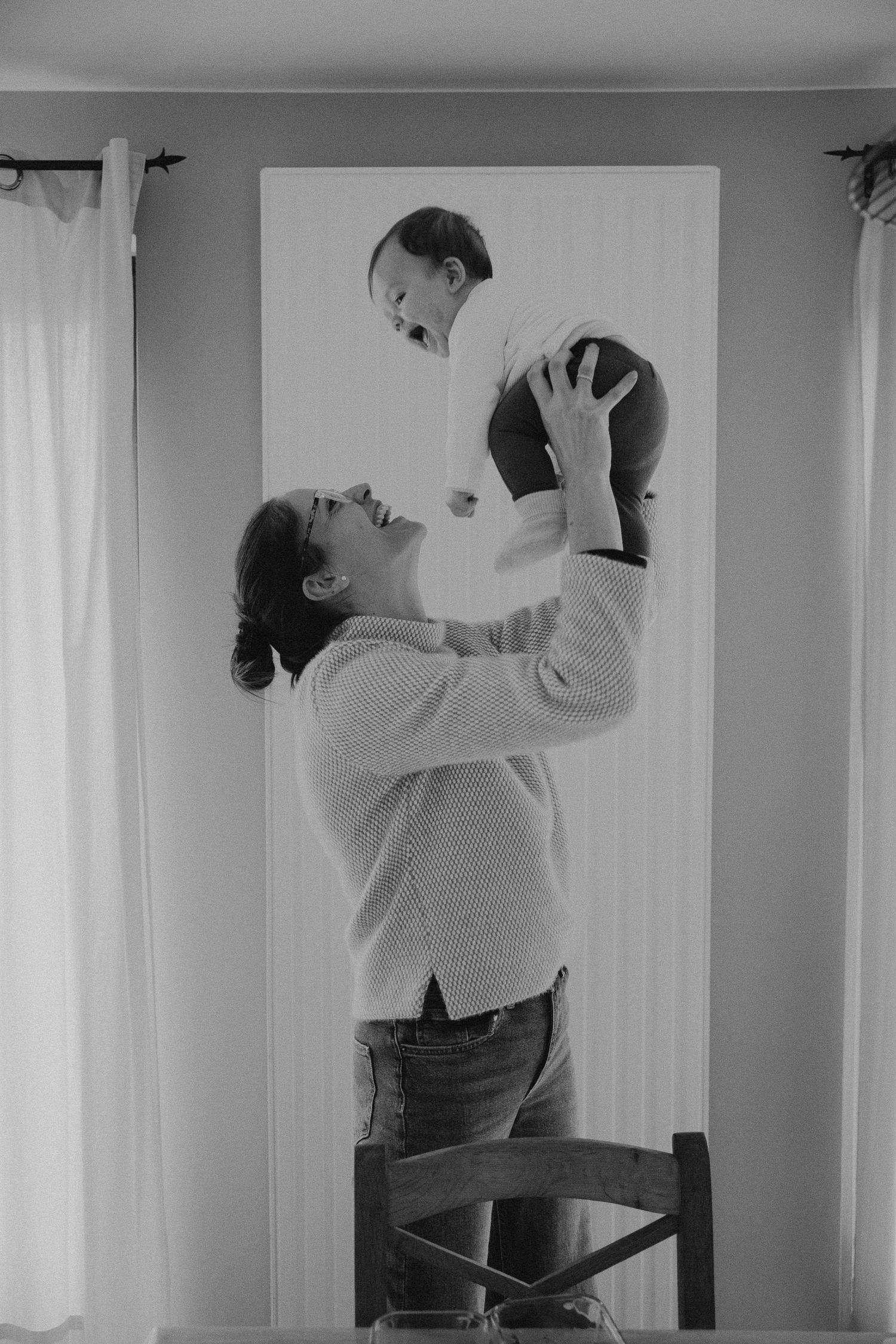 Black and white photo of a mother lifting her laughing baby above her head while standing on a chair indoors.
