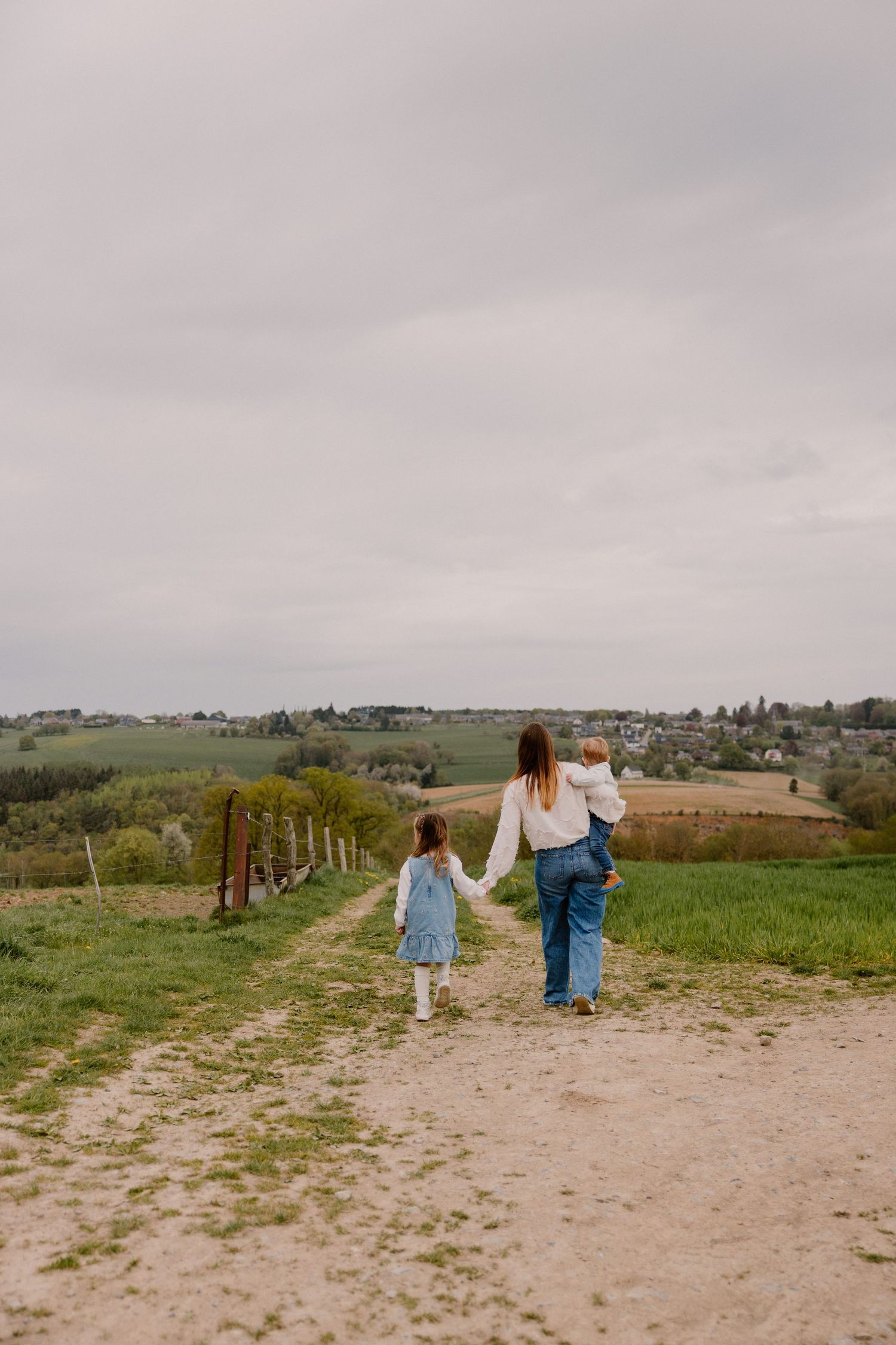 Mother walking with two young children on a rural dirt path surrounded by green fields under a cloudy sky.