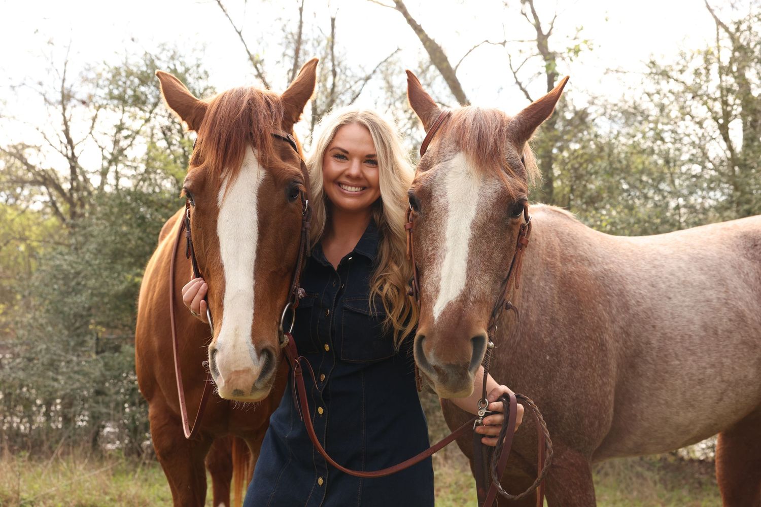 Smiling blonde woman standing between two chestnut horses in a wooded outdoor setting.