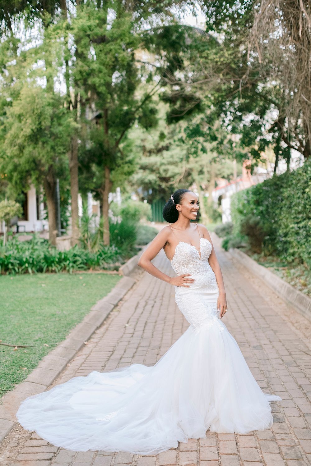 Stunning bride in strapless beaded mermaid gown with long train poses on brick pathway in garden.