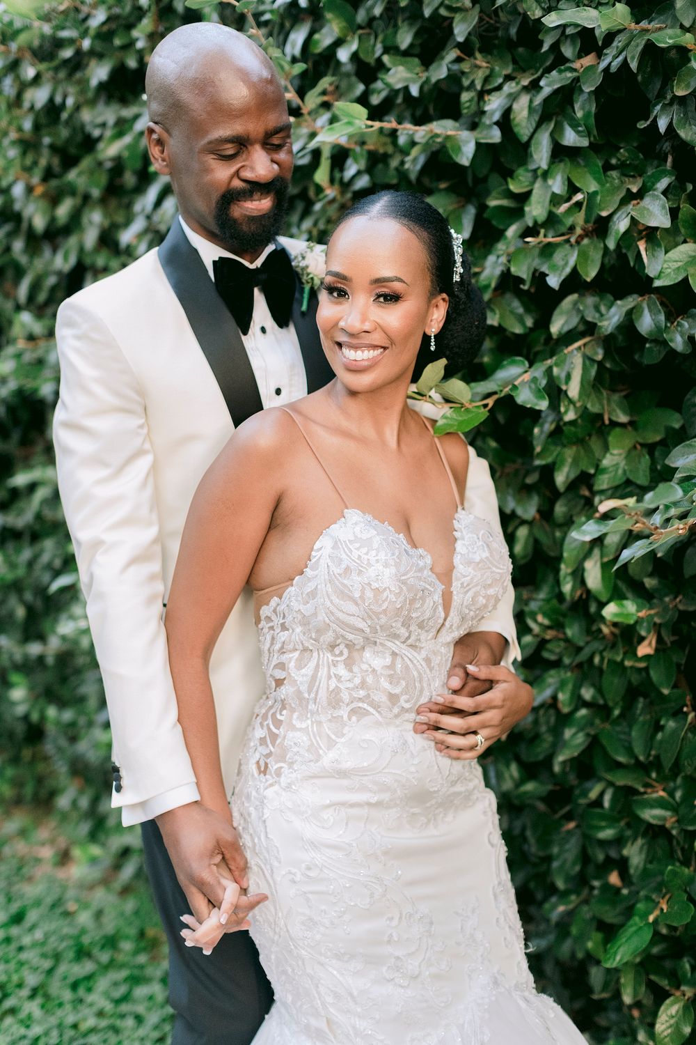 Groom in white tuxedo embraces smiling bride in beaded gown against lush green ivy-covered wall.