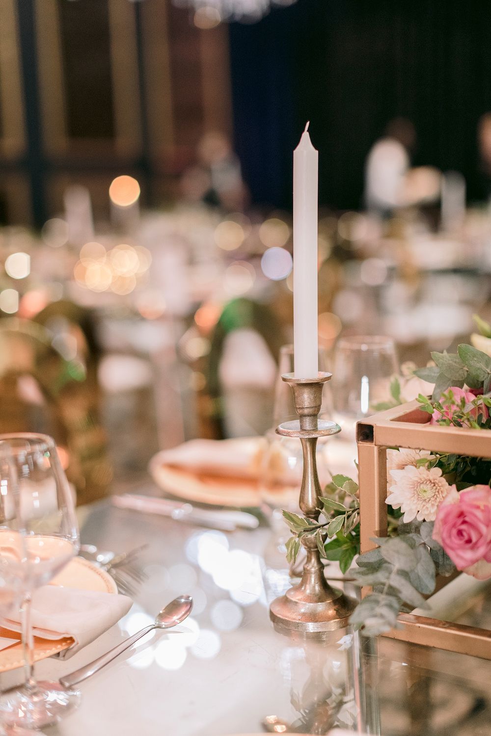 Elegant wedding reception table with silver candlestick, white taper candle, and pink floral centerpiece on mirrored surface.