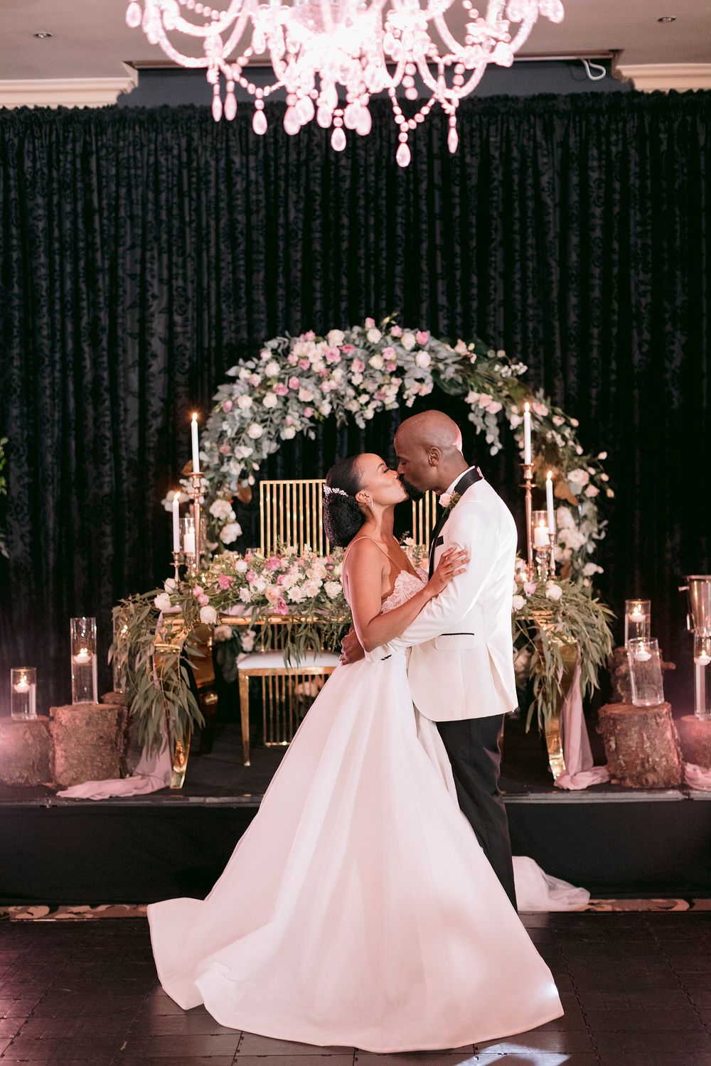 Bride and groom sharing first dance under floral arch with candles and lush greenery in elegant ballroom.