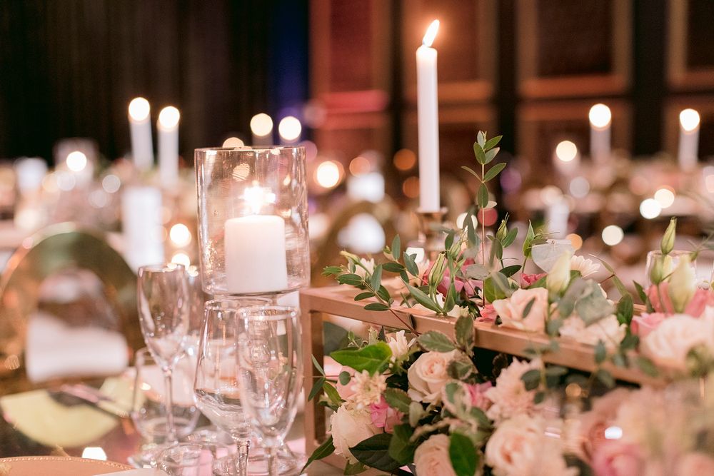 Elegant wedding reception table with white pillar candles, pink roses, and crystal glassware in a candlelit ballroom.