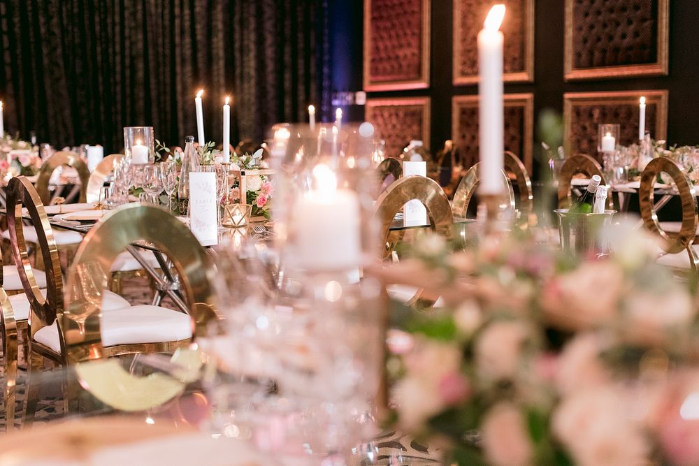 Softly lit wedding banquet hall with floral centerpieces, tall taper candles, and gold chairs creating an elegant atmosphere.