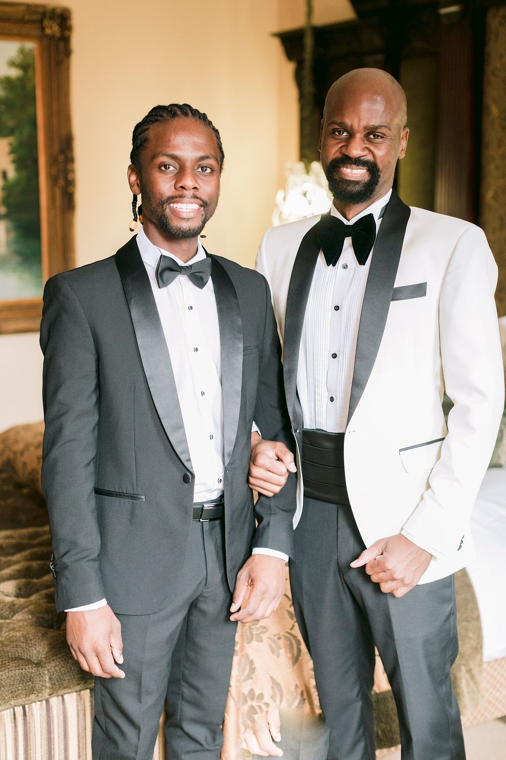 Two groomsmen in formal black and white tuxedos smile together at a wedding preparation.