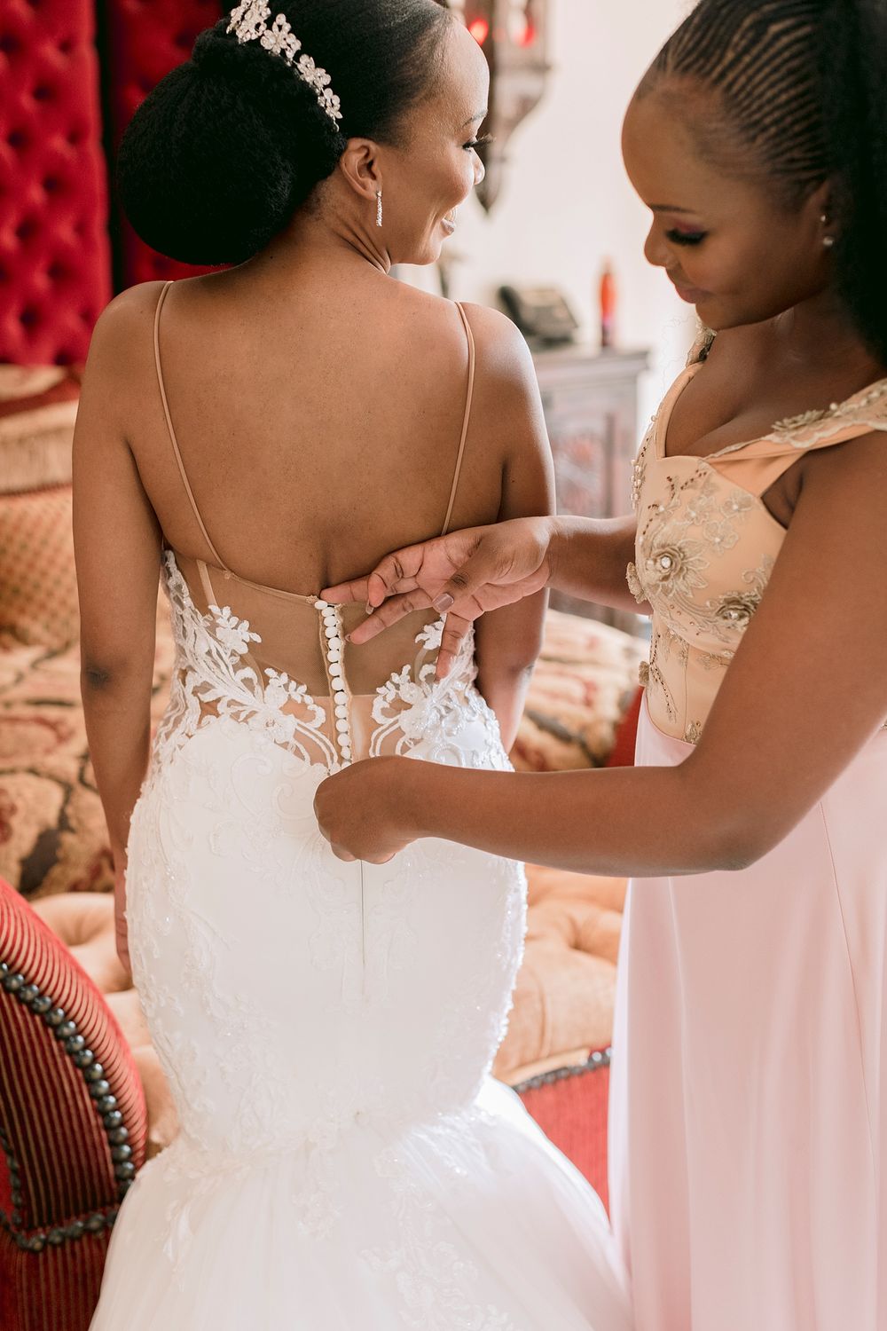 A bridesmaid in pink helps button the back of a bride's white lace wedding gown.