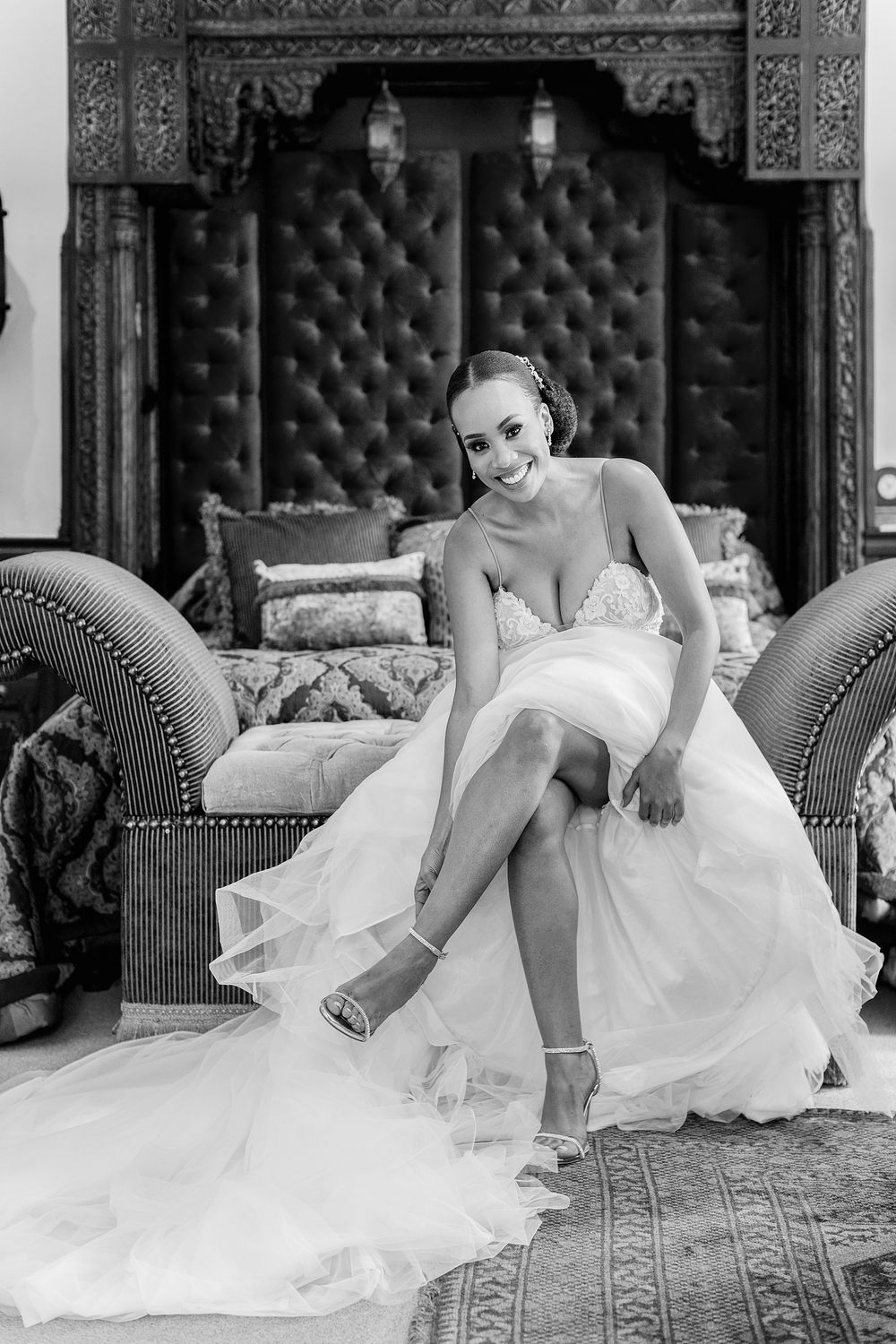 Black and white photo of a radiant bride in a lace gown putting on her wedding shoes.