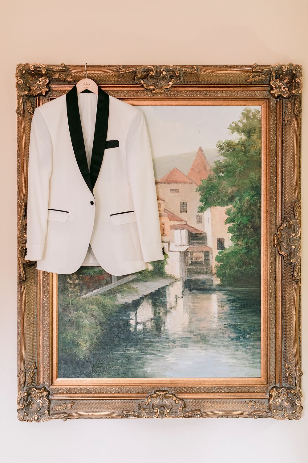 White tuxedo jacket with black lapels hanging on ornate gold-framed oil painting of European waterway scene.