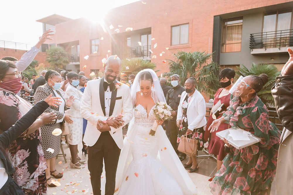 Joyful newlyweds walk through confetti shower outdoors as wedding guests celebrate in sunlight.