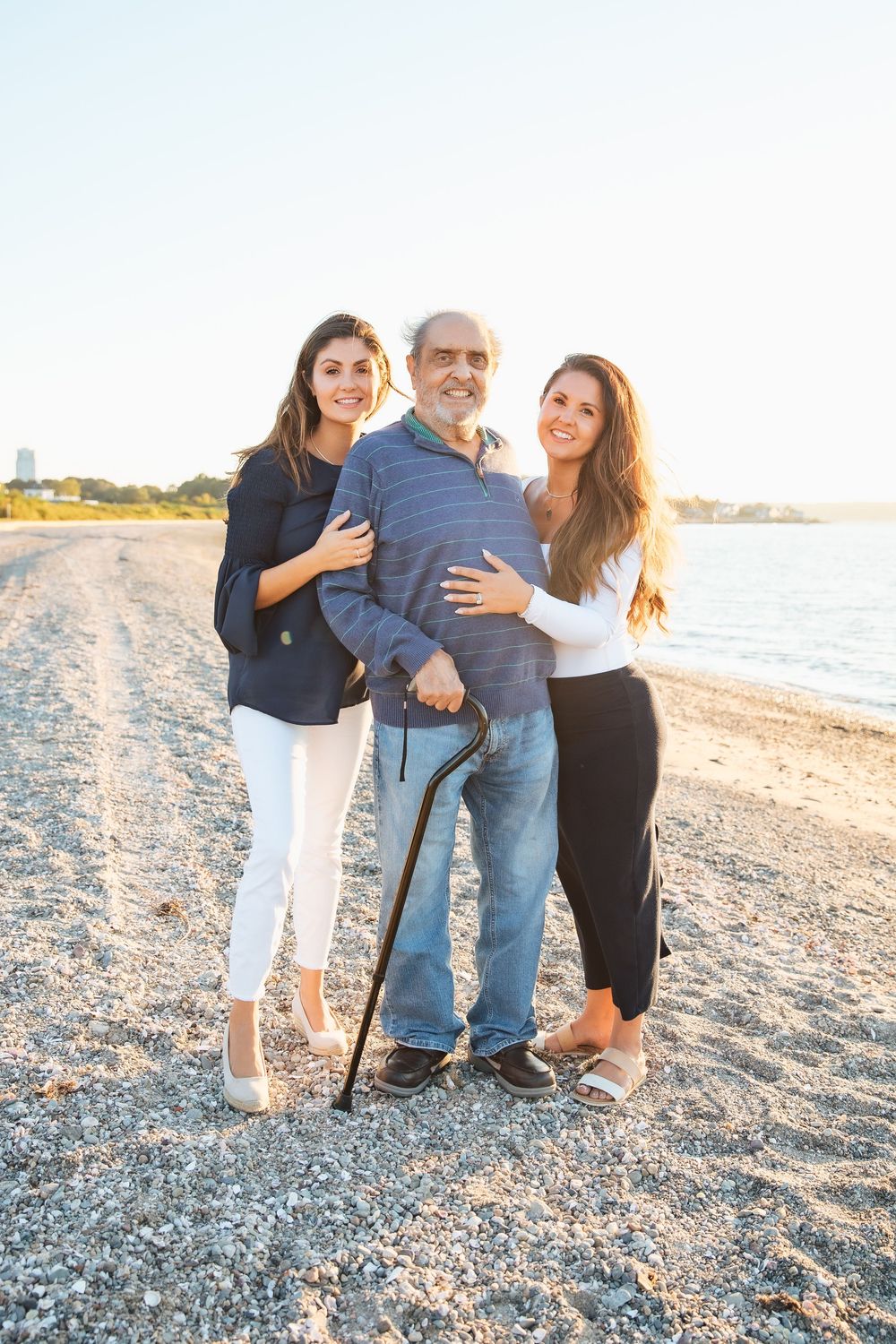 Elderly man with cane poses with two young women on a sunny beach, all smiling warmly together. extended family photoshoot Portsmouth RI