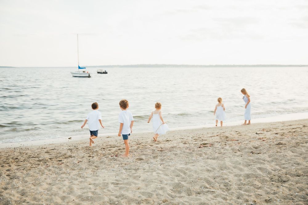 Five children in white clothing running and playing on a sandy beach with a sailboat in the ocean background. . Extended Family photoshoot