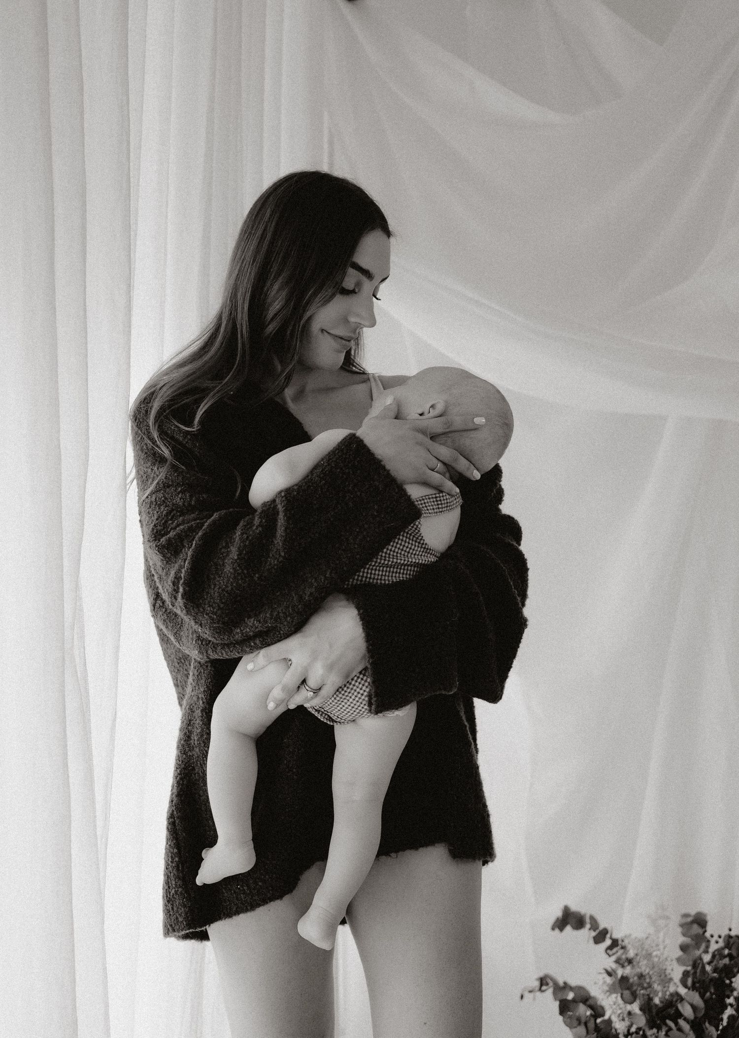 Black and white photo of a young mother tenderly cradling her newborn baby near sheer white curtains.