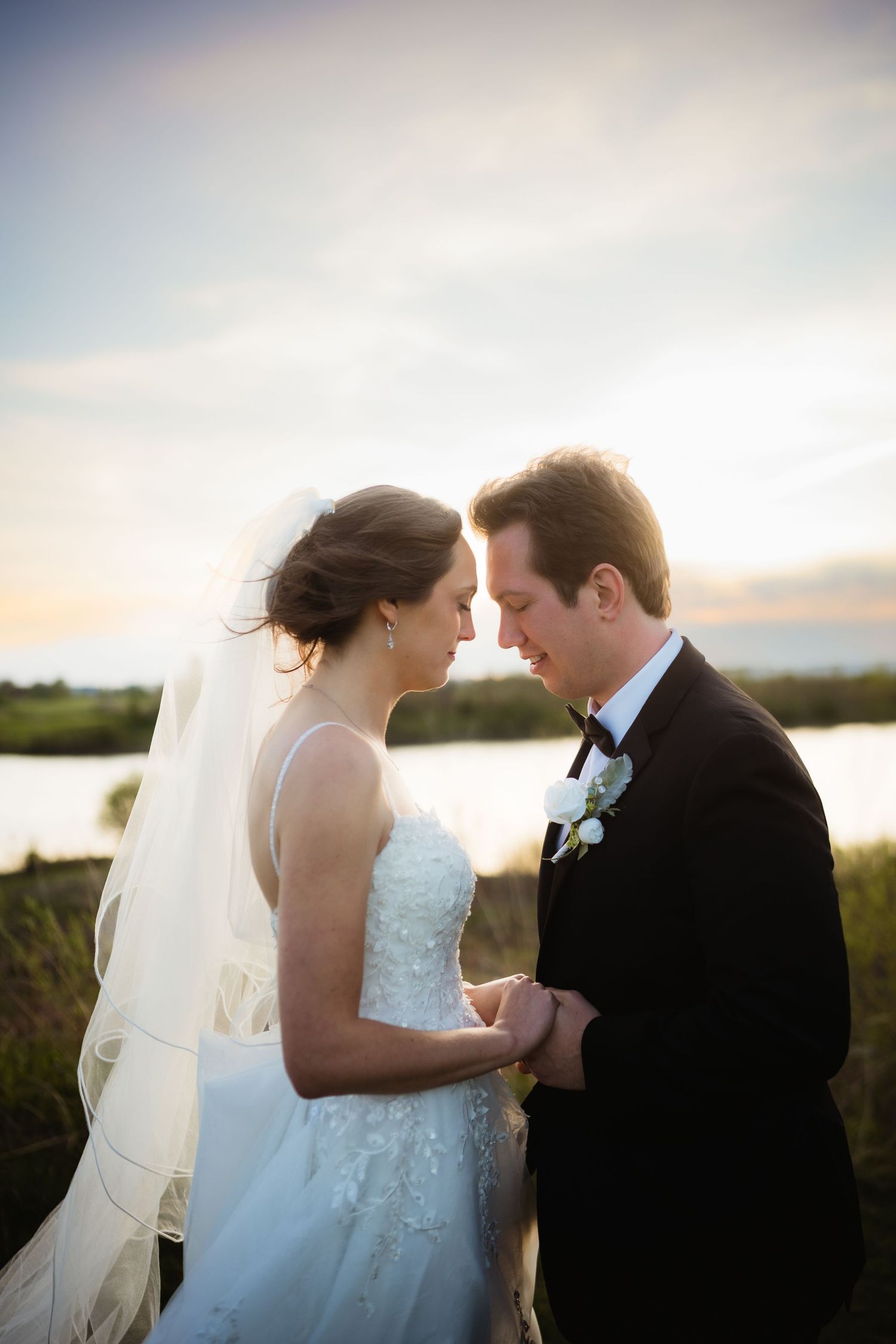 Bride and groom touching foreheads at sunset near a serene waterway, bride in lace gown with veil, groom in black tuxedo.