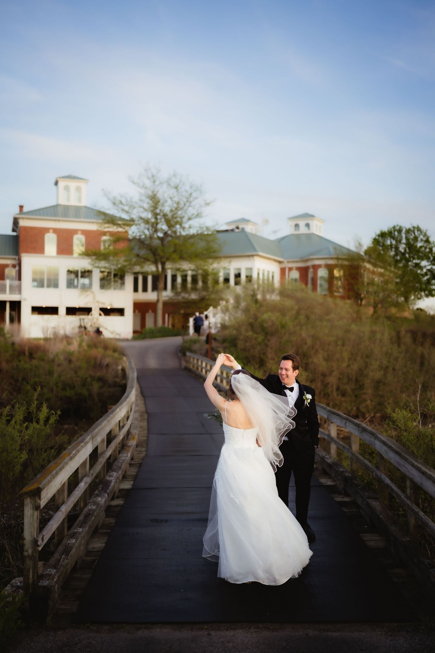 Bride and groom dancing on a wooden bridge at sunset, with a brick wedding venue in the background.