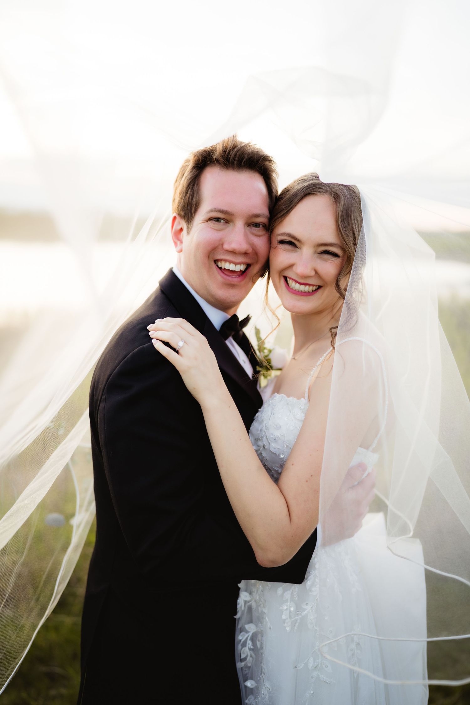 Smiling bride and groom embrace outdoors, bride wearing lace gown and veil flowing in the breeze on their wedding day.
