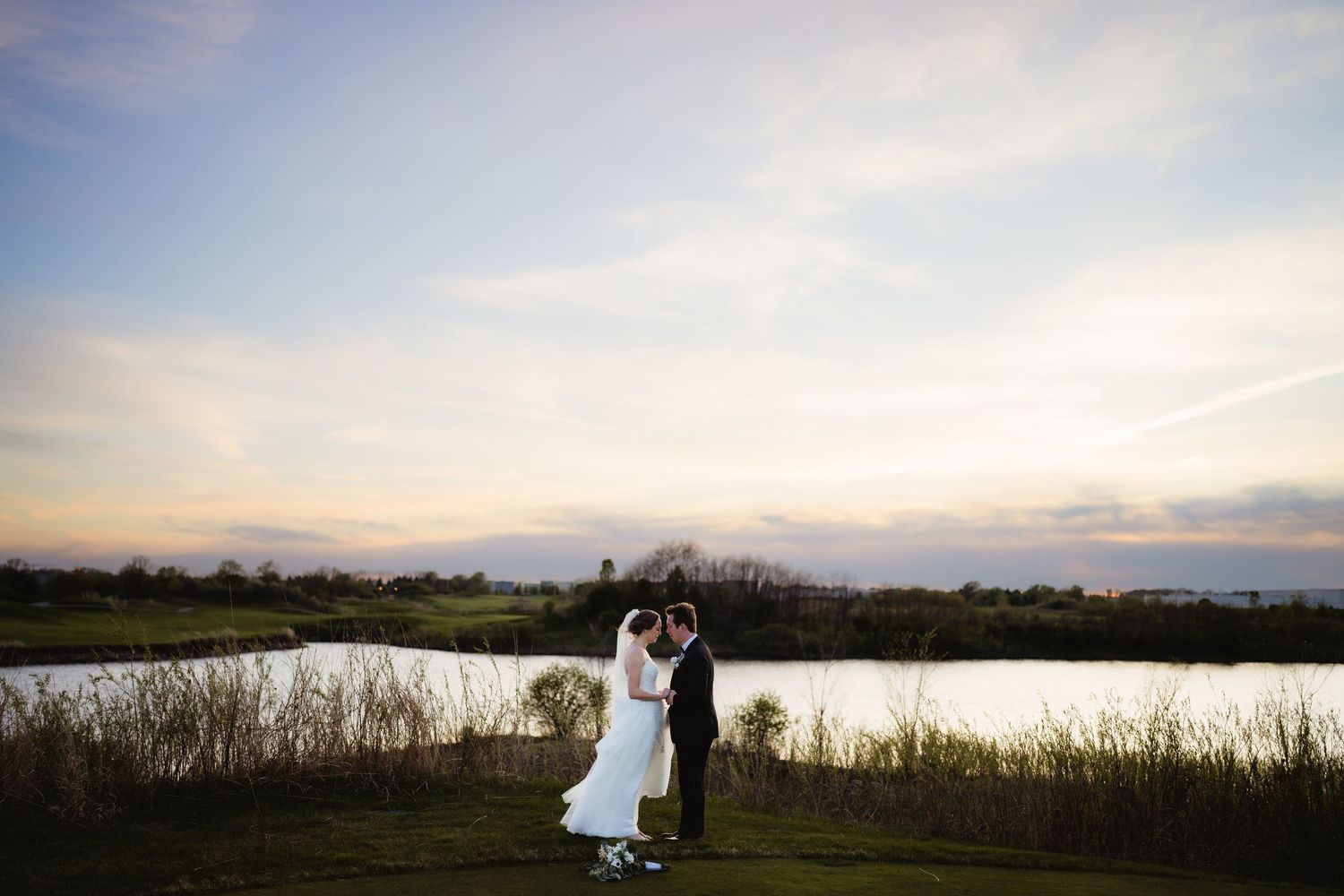 Bride and groom share a romantic kiss at sunset by a serene lake, with golden sky and reeds in the background.