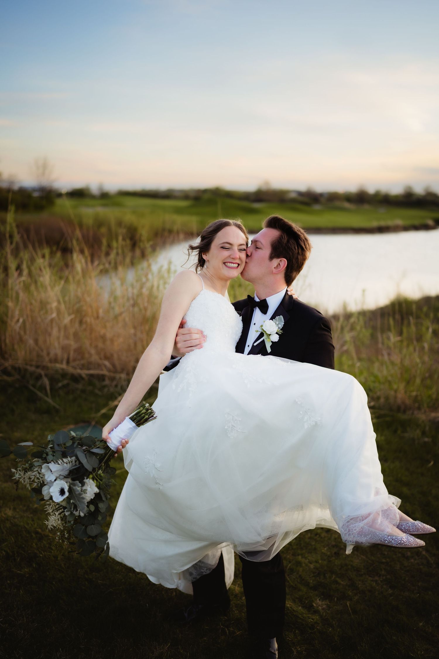 Groom in black tuxedo carries smiling bride in white gown outdoors near pond at sunset.