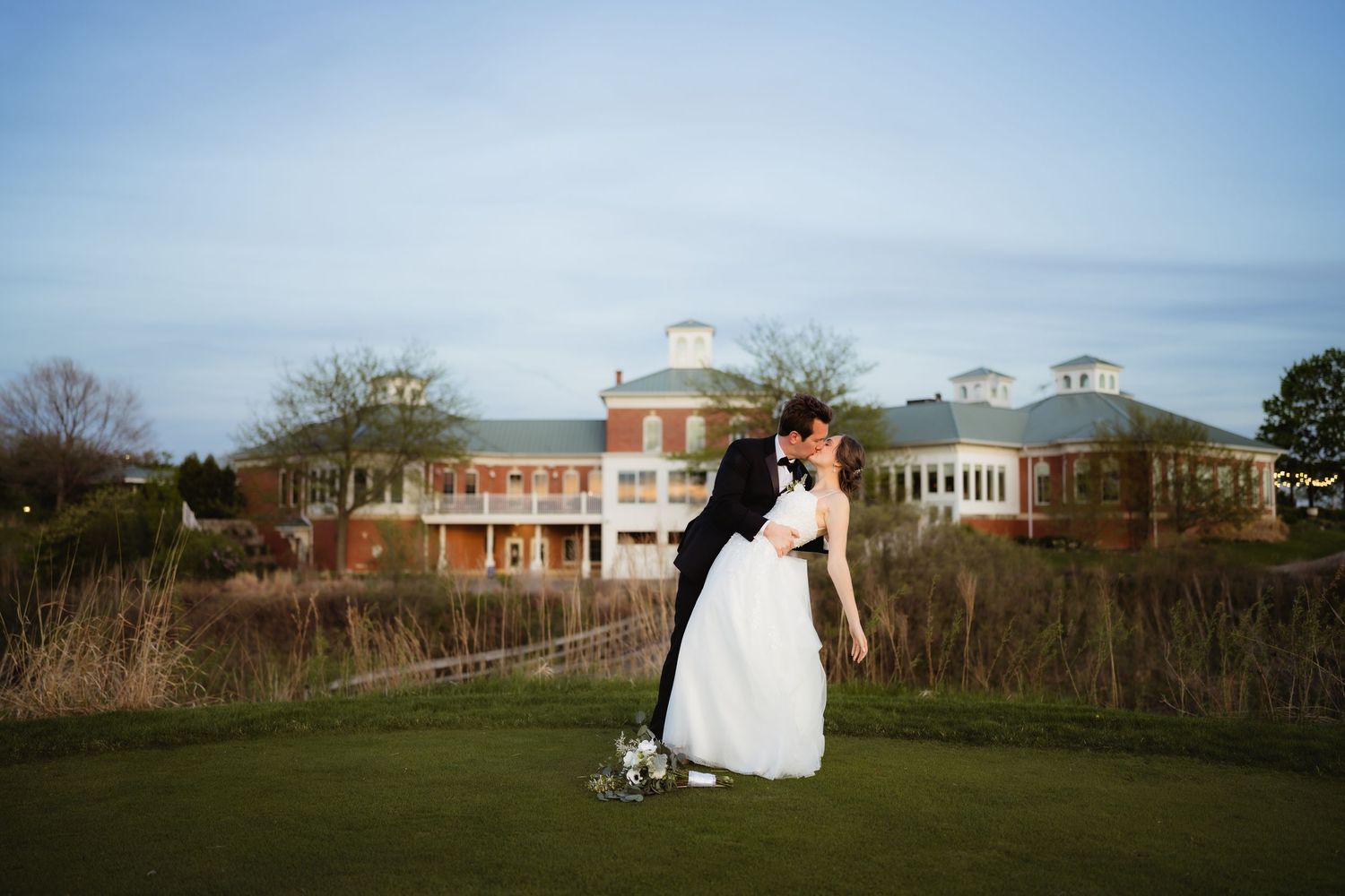 Bride and groom share a romantic kiss on a green lawn with an elegant brick wedding venue in the background.