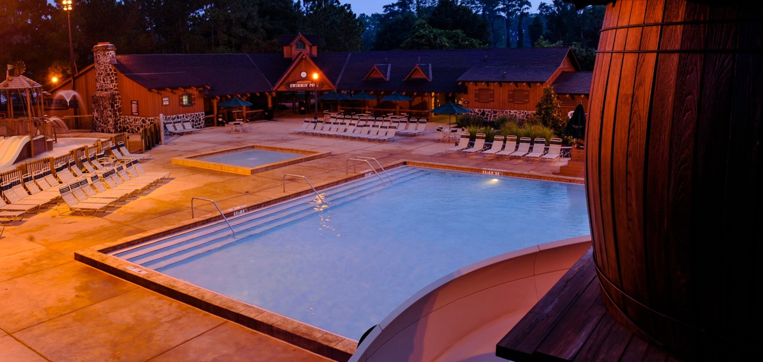 Illuminated swimming pool area at night with lounge chairs and wooden barrel structures.